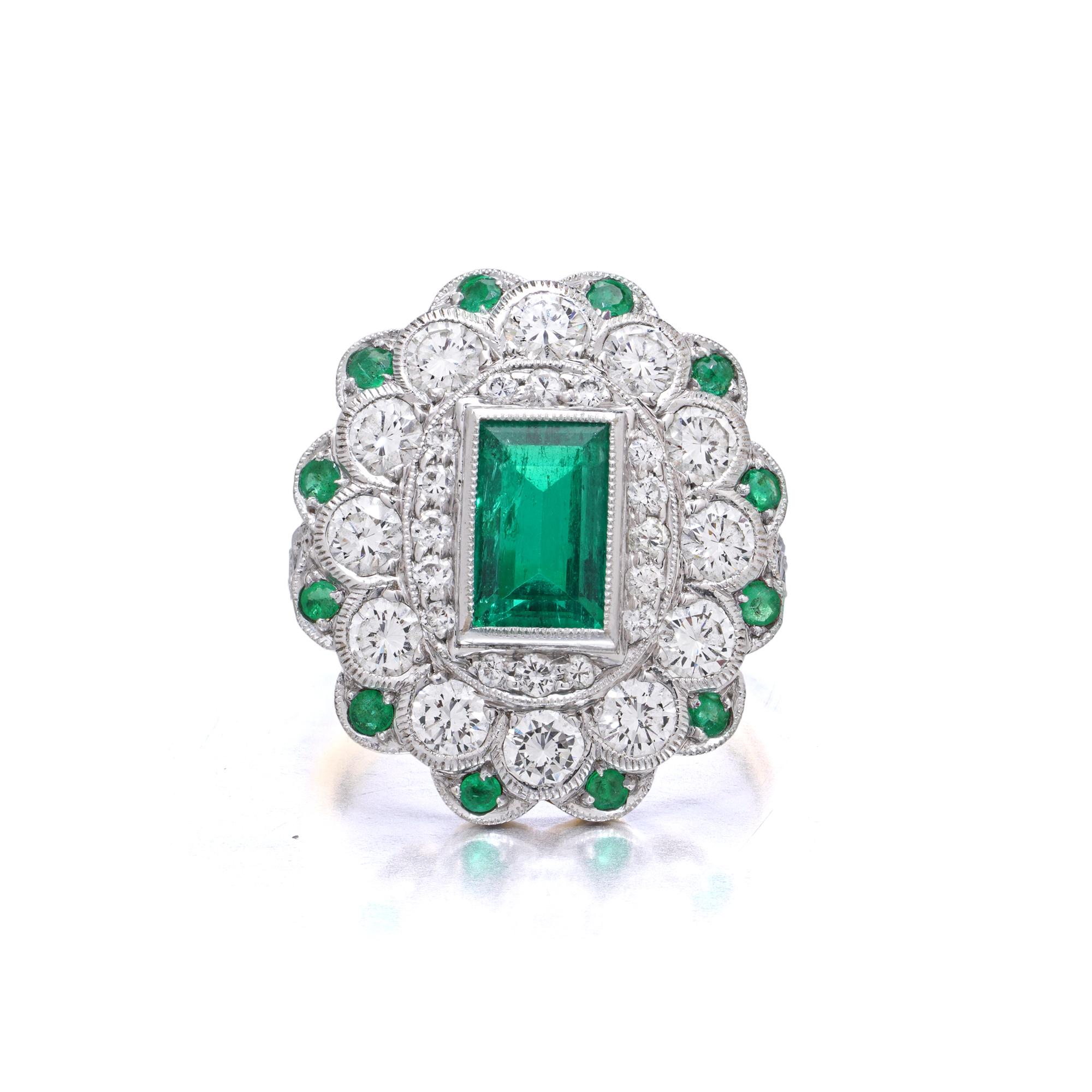 Un vibrante y elegante anillo en racimo de oro blanco de 18 quilates engastado con una vívida esmeralda rectangular en su centro, enmarcada por un contorno de diamantes talla brillante y otro borde de esmeraldas redondas facetadas. Con una silueta