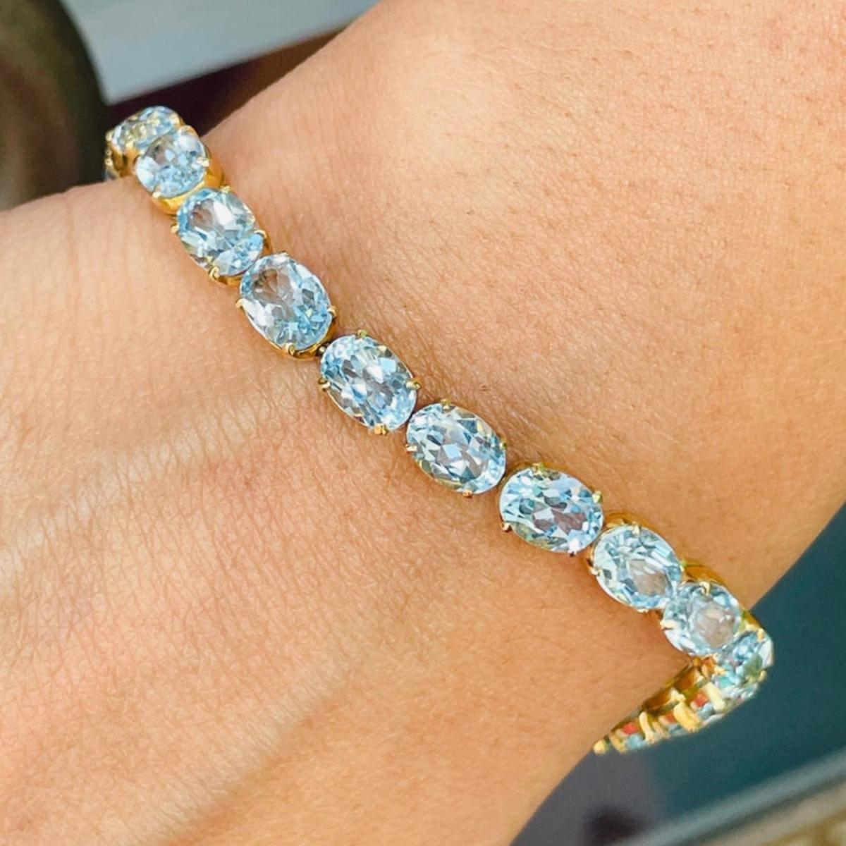 Ce bracelet de tennis en or 18 carats orné de topazes bleues étincelantes met en valeur 25 topazes bleues naturelles d'un poids de 28,5 carats qui scintillent à l'infini. Il mesure 7.5 pouces de long. 
La topaze bleue aide à améliorer la