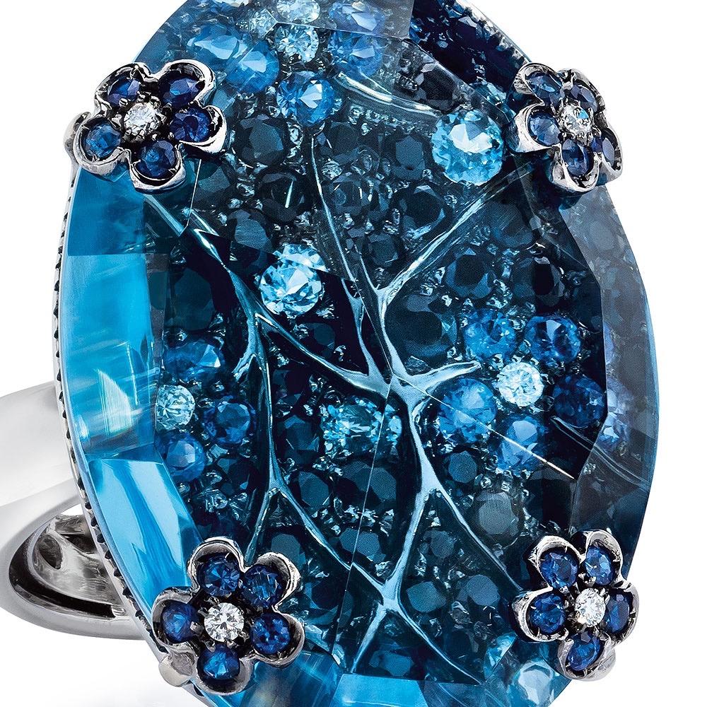 This stunning rich blue oval-shaped ring is composed of .80 carats of blue sapphire cluster flowers surrounded by 2.40 carats of black sapphires and .25 carats of aqua. This detailed piece is overlayed with a 28 carat blue topaz that magnifies the