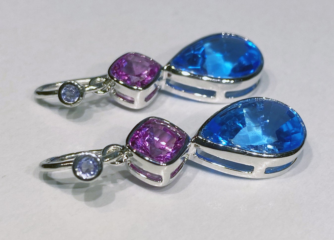 18kt White Gold  Earrings set with Tanzanite, Blue Topaz & Cultured Pink Sapphires. 18kt White Gold weight is 5.3 Grams, Earring French Hooks are Silver, body of earings are 18kt White Gold. Two small Tanzanite rounds accent the silver French Hooks