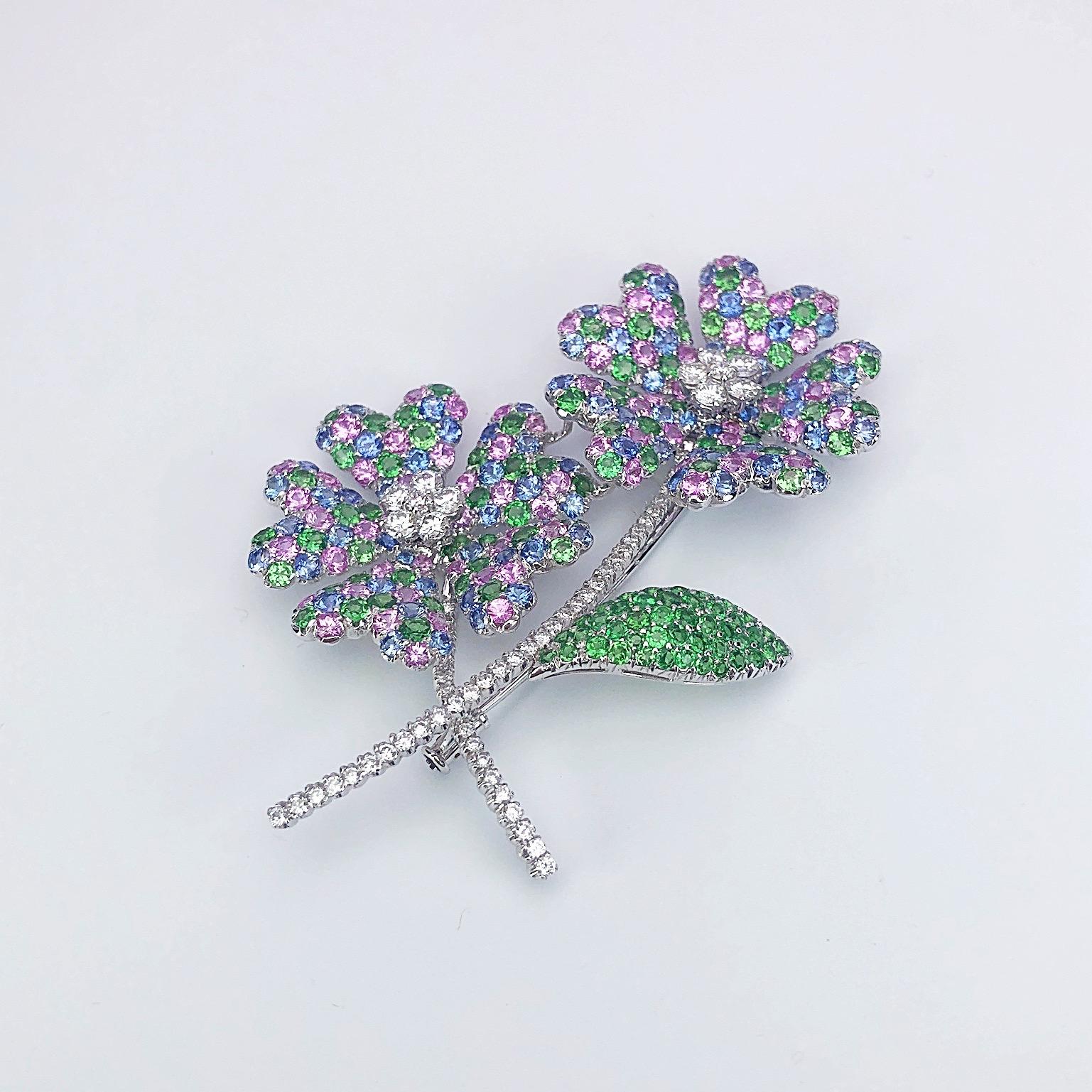 18 karat white gold brooch designed with two pastel hued flowers. The petals are set with round  pink and blue sapphires and green tsavorites. The center of each flower and their stems are set with round brilliant diamonds. One leaf is set with