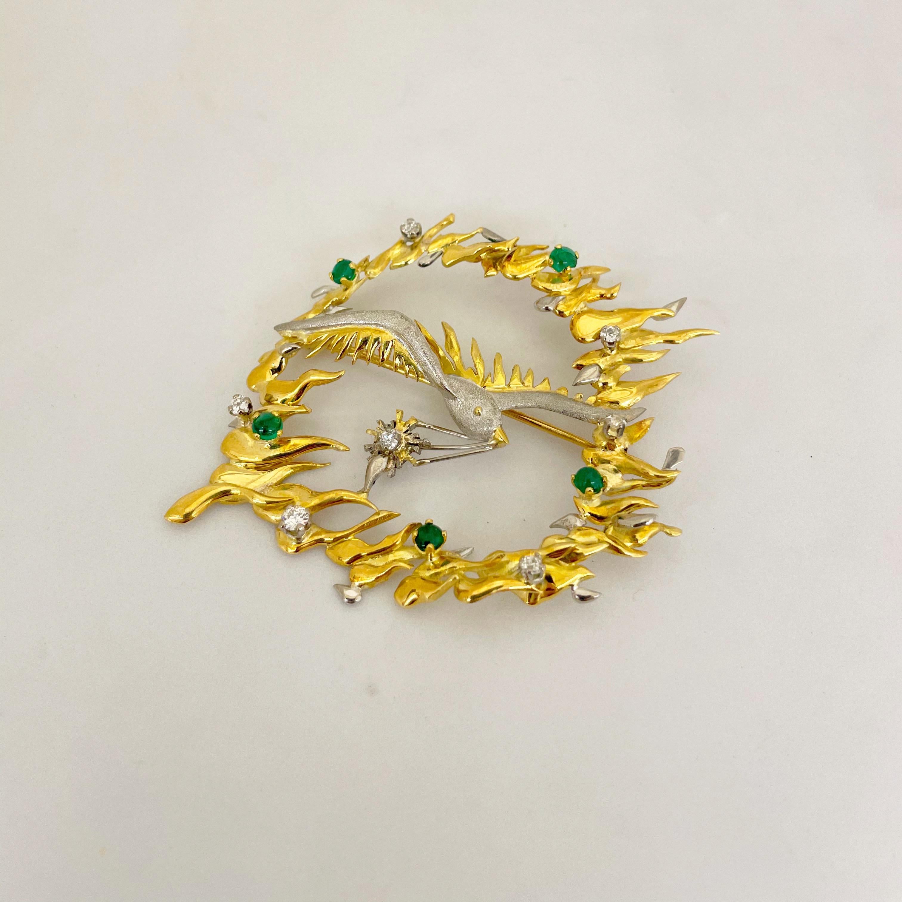 Vintage 18 karat gold brooch. The brooch is designed with a bird motif in a white gold matte finish, with shiny yellow gold for the feathers. The bird sits in a ring of fire which has been accented with round brilliant diamonds and cabochon emeralds