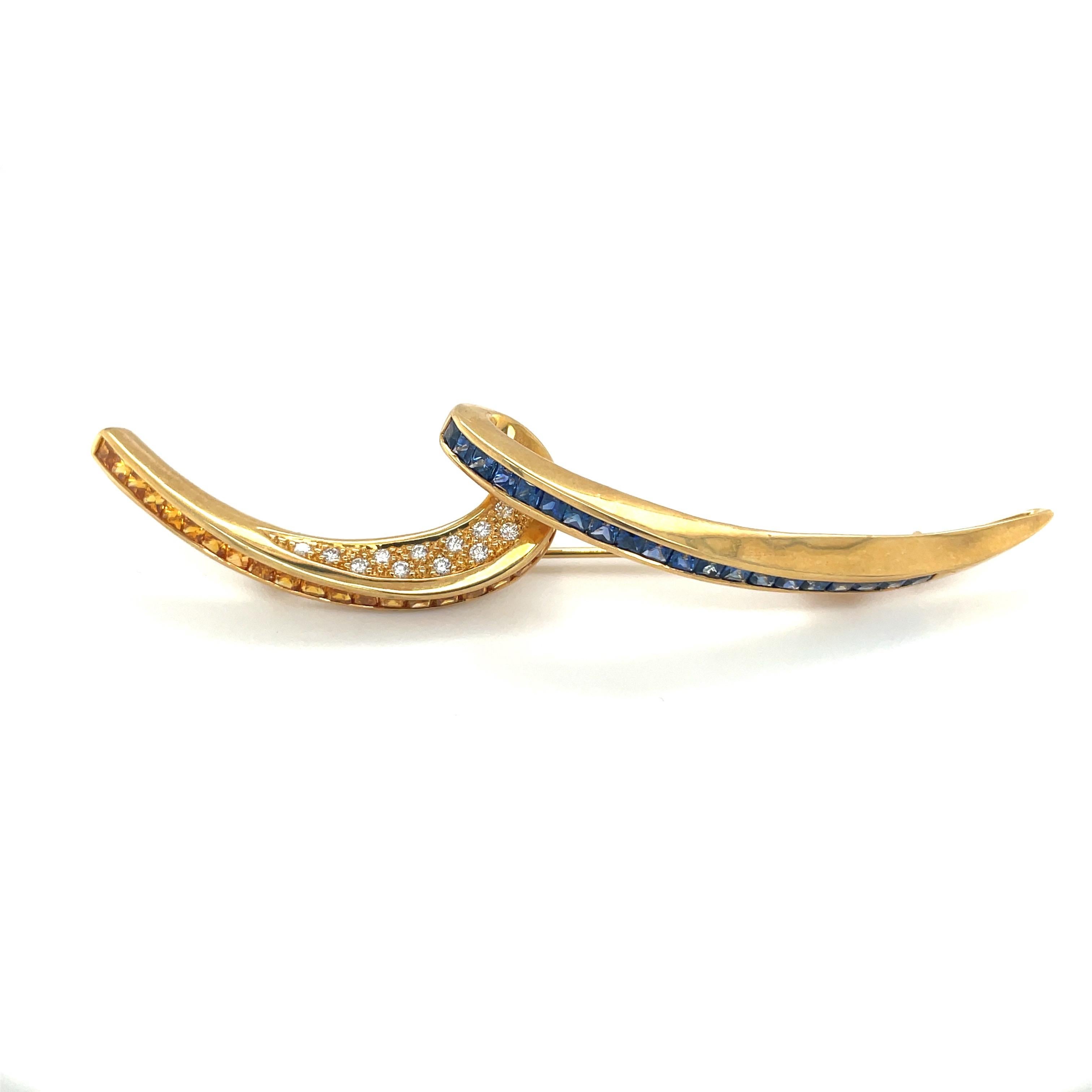 Cellini Jewelers Designed as a fluid ribbon, this 18 karat yellow gold brooch is invisibly set with 3.10 carats of princess cut blue and yellow sapphires . Round diamonds 0.20 carats accent the inside of the ribbon. The elongated brooch measures