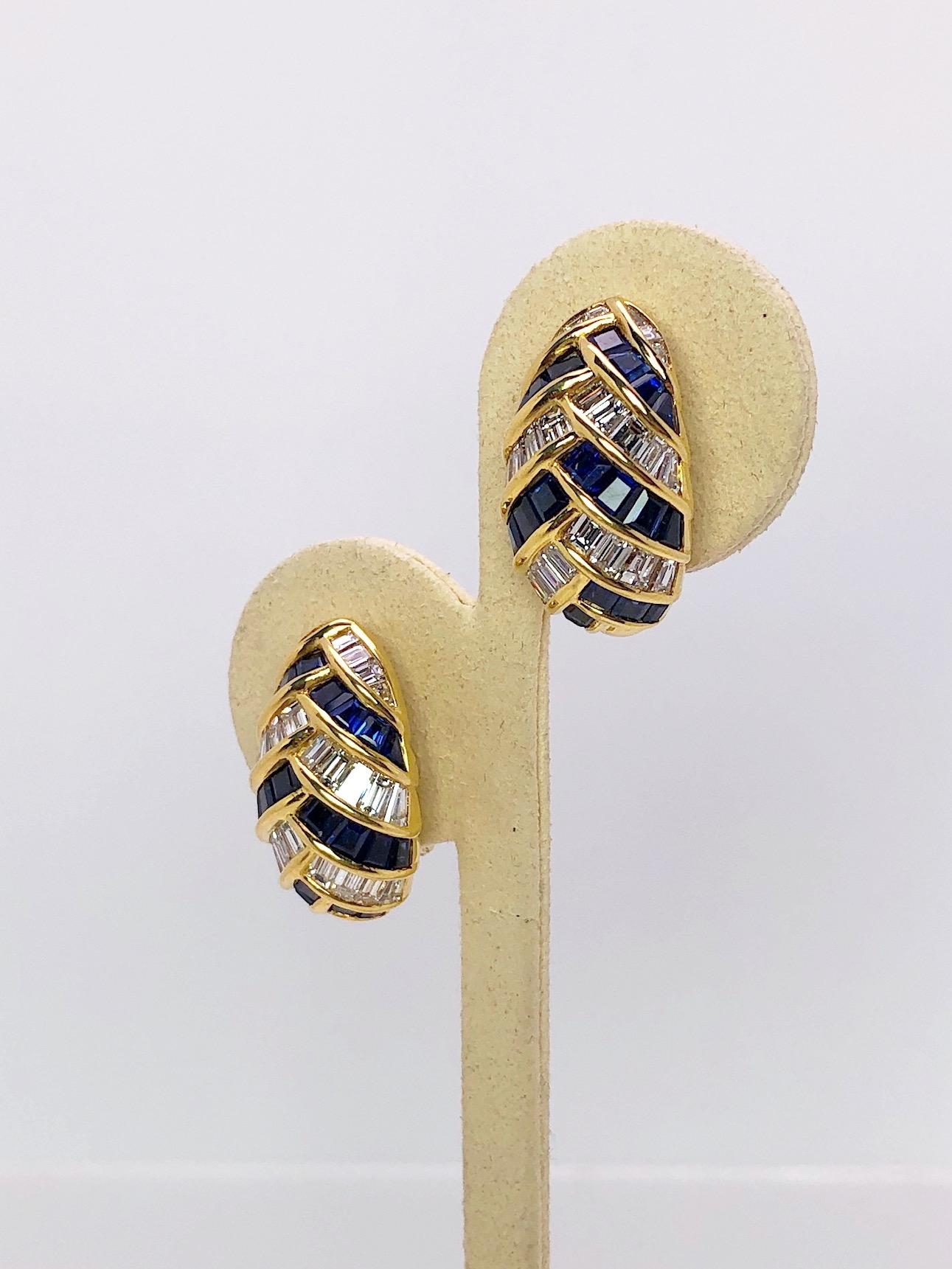 A classic oval shaped 18 karat yellow gold earring is impeccably set with Baguette Cut Diamonds and Blue Sapphires. The stones are set in an alternating weaved pattern with gold bezels. The earrings measure 1