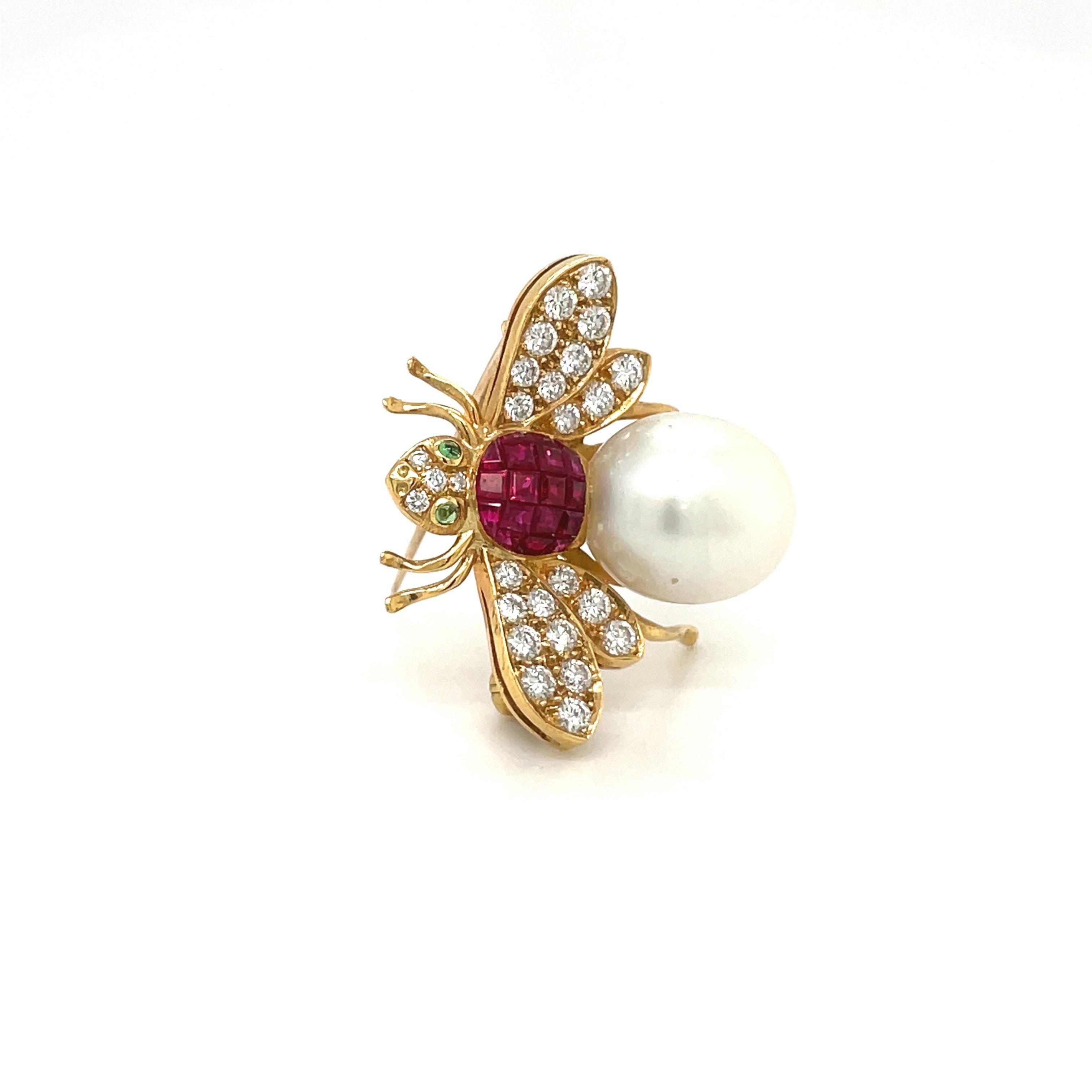 18 karat yellow gold bee brooch. This lovely brooch is beautifully designed with a body of invisibly set rubies and a South Sea pearl. The wings and head are composed of round brilliant diamonds. Two cabochon emeralds are set as the eyes. The bee