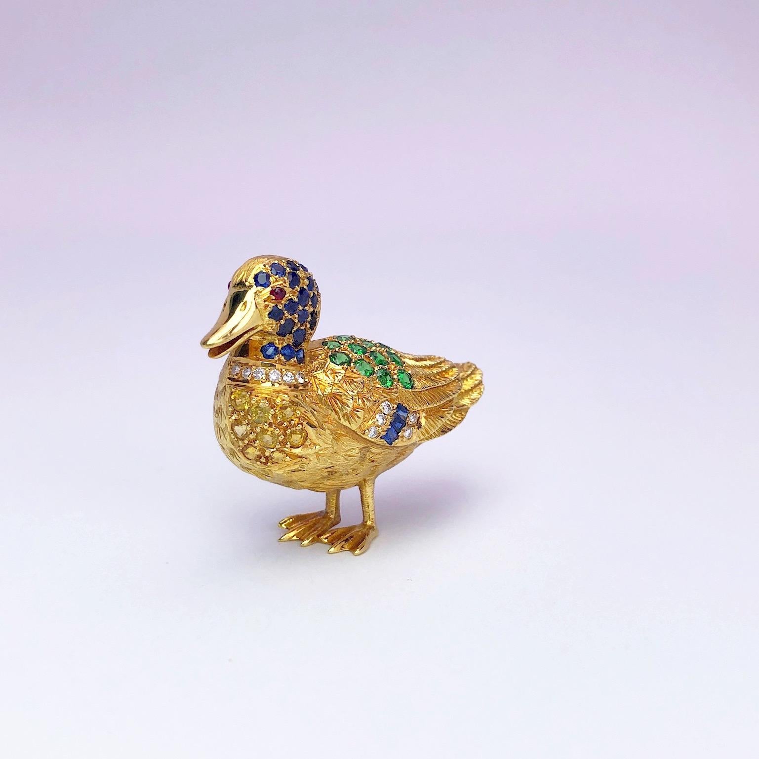 This 18 karat engraved yellow gold duck brooch is set with  1.17 carats of blue and yellow sapphires, 0.65 carats of tsavorites,  0.10 carats of diamonds , and a ruby cabochon. 
The duck measures 1.25