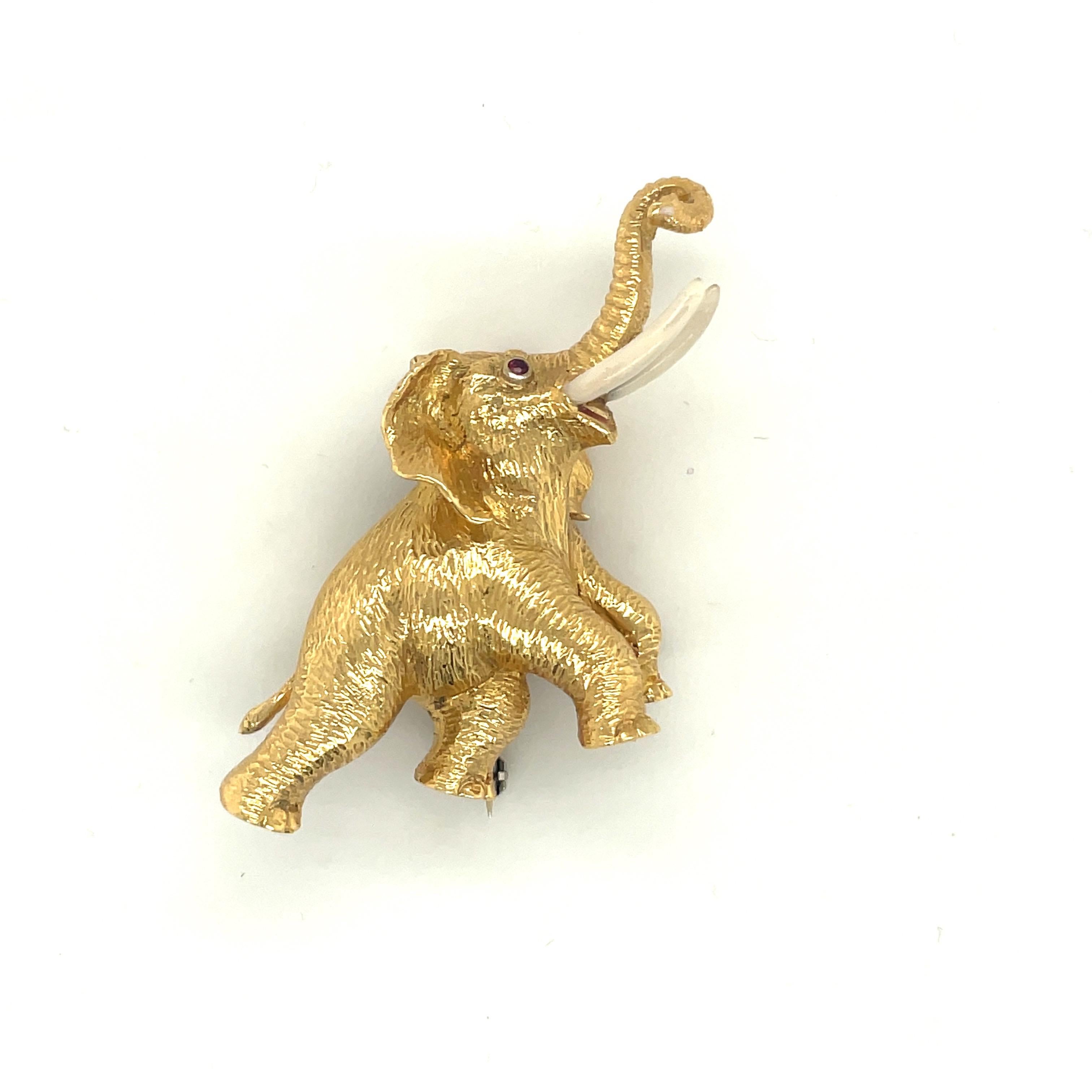This playful elephant brooch is designed in a sandblasted 18 karat yellow gold finish. His eyes are set with  round rubies and his tusks are a white enamel .The elephant's trunk is lifted up, which represents good luck!
