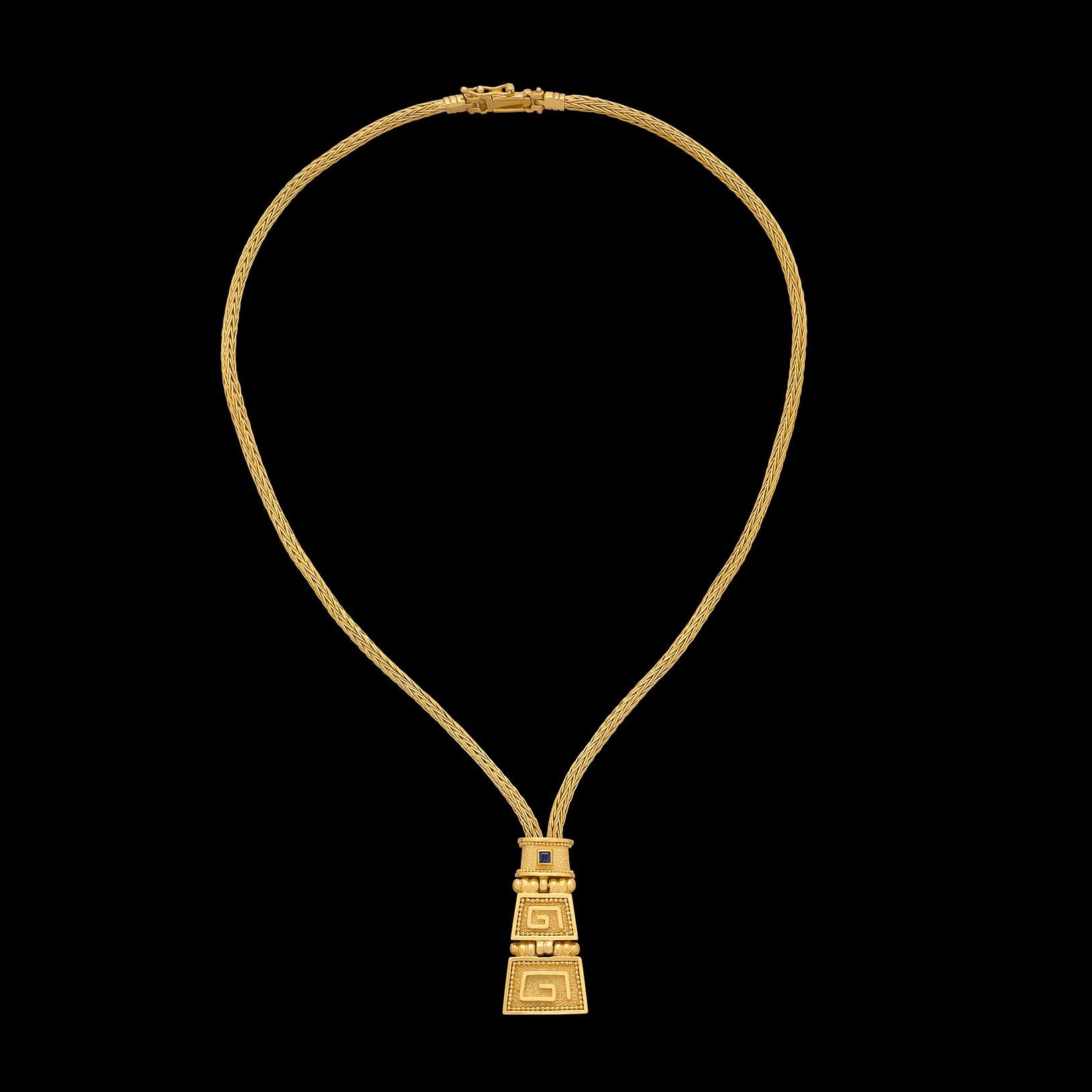 A true head-turner! This 18 karat yellow gold necklace features a gold twist chain with a Greek pattern inspired pendant suspended from it. Bezel set within the gold pendant is a single blue sapphire, adding just the perfect pop of color. The gold