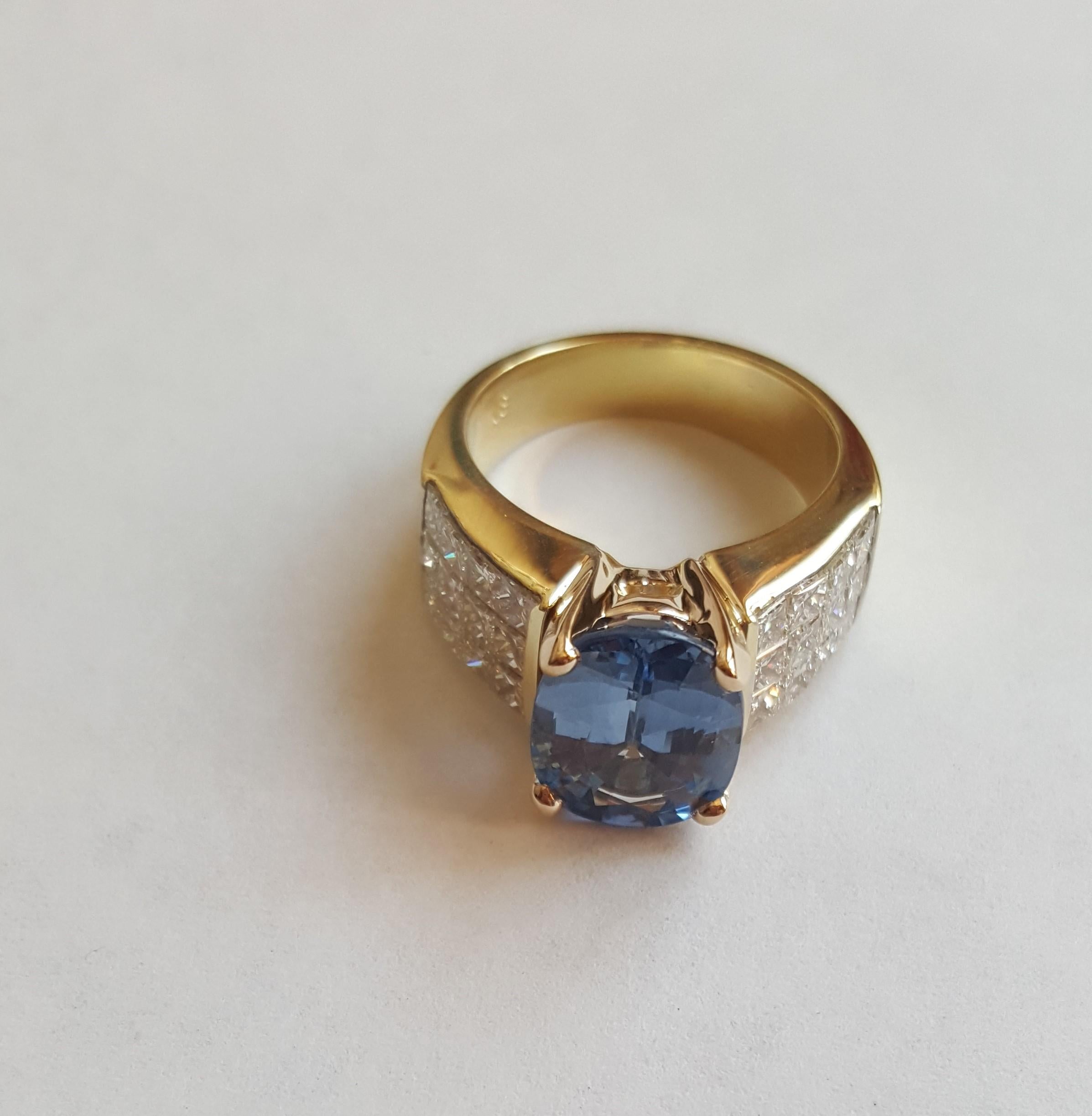 18kt Yellow Gold Oval 4.45ct Blue Sapphire Ring with 24 Princess Cut Invisible Set Diamonds. The blue sapphire is 11.4mm x 8.6 mm in diameter and has a nice medium blue cornflower color, a very clean stone, set in a 4-prong basket style setting. 