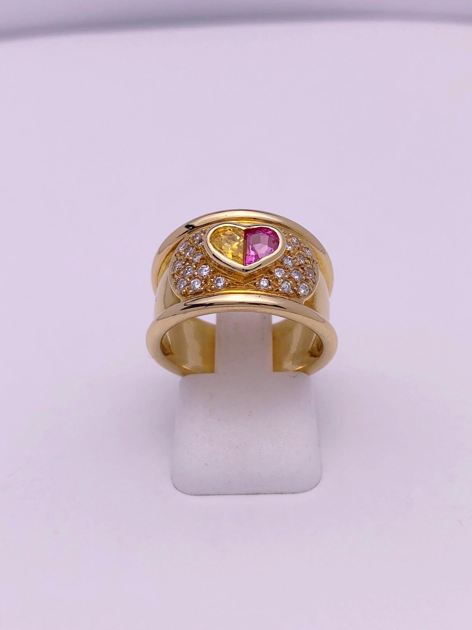 This band ring centers an 18 karat yellow gold heart bezel set with two sapphires. One yellow sapphire and one pink sapphire set side by side form the heart. The heart sits amid a pave diamond section. Two raised bezels form the outer portions of
