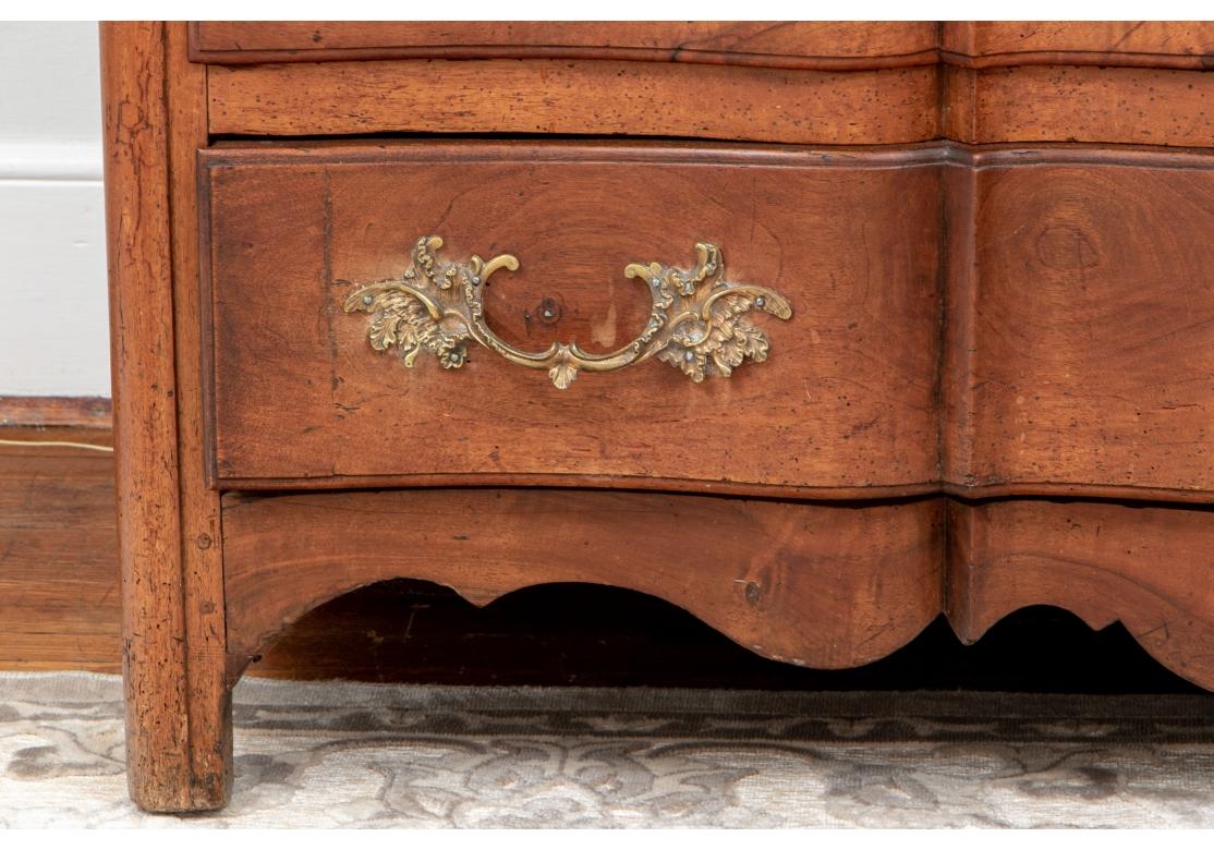 A French antique chest with particularly fine form in original condition. An antique country French three drawer chest with rococo brass pulls and escutcheons, one key, planked top and back, raised panel sides, serpentine front, scalloped apron and