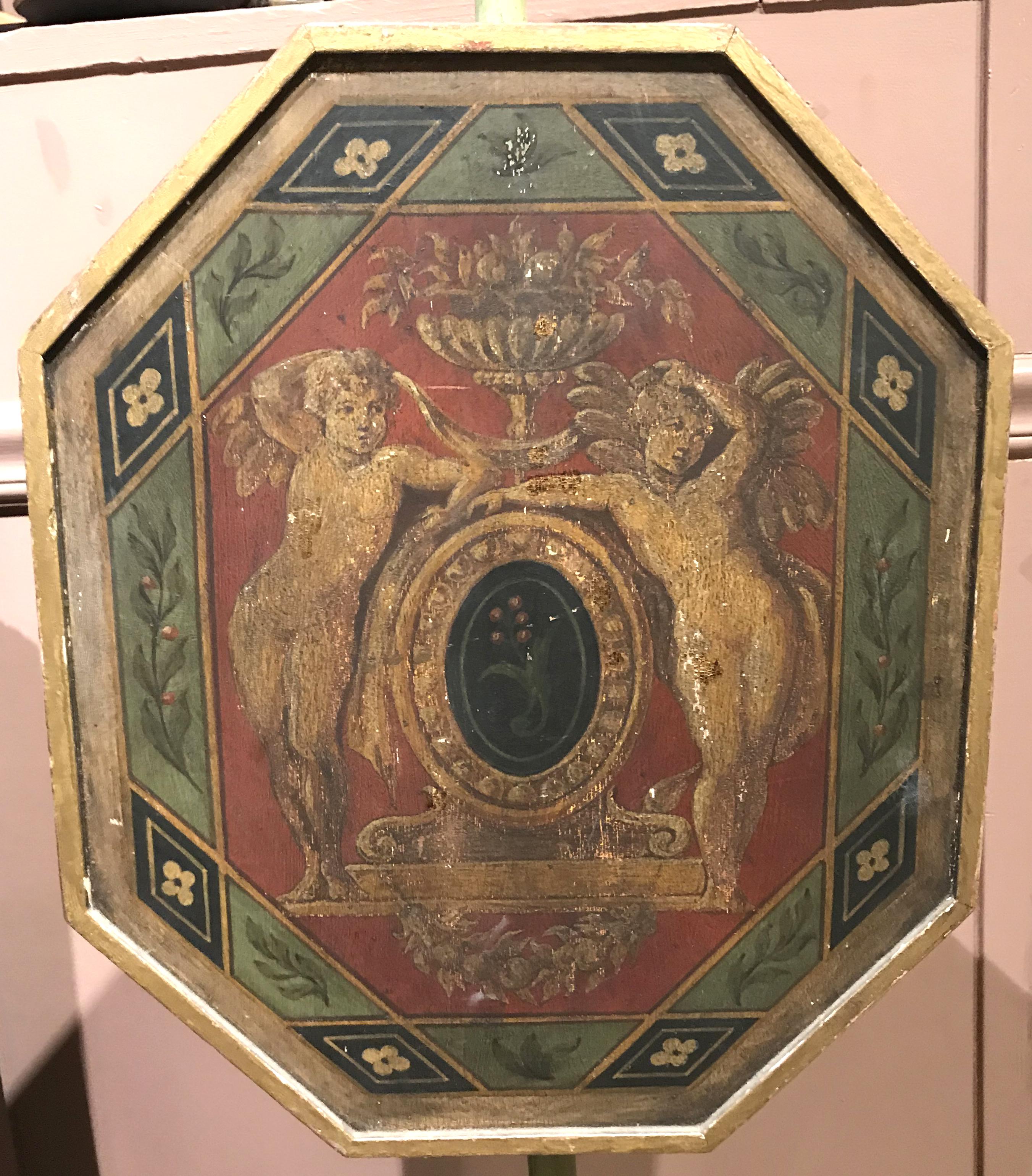 A wooden light green painted pole screen with octagonal adjustable panel with early polychrome hand painted cherub decoration on an urn form tripod base with gilt highlights. English in origin, dating to the late 18th or early 19th century. Good