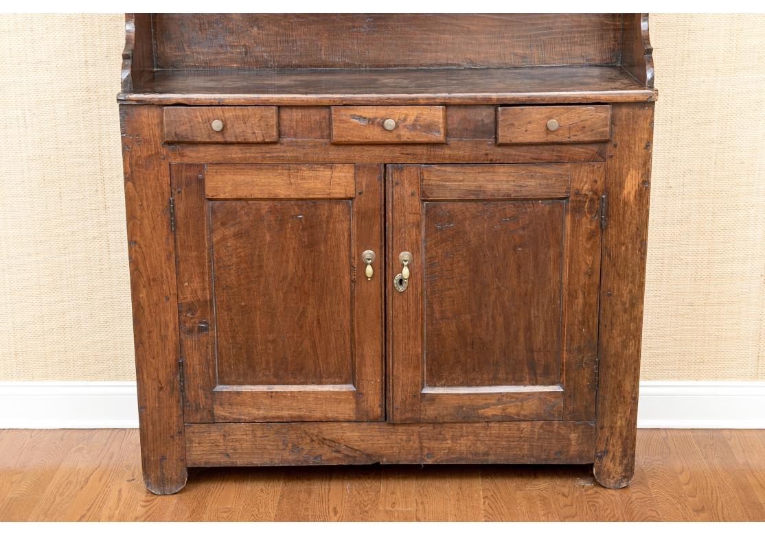 An Antique English Hutch with fine age patina, soft timeworn hand and fantastic traditional form.  A nicely patinated oak cupboard with scalloped plate racks having three short drawers over double cabinet doors with brass teardrop pulls.Lacking a