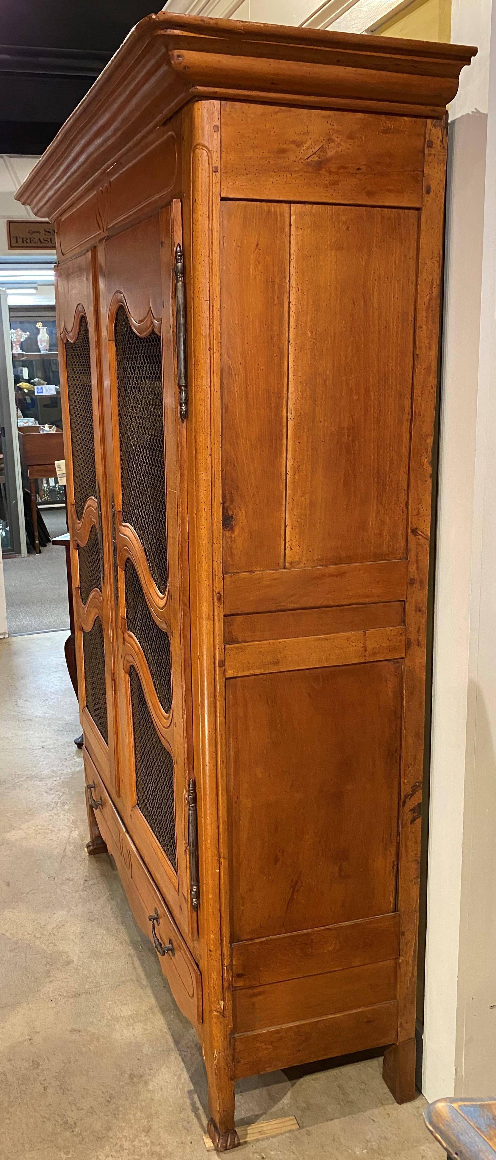 A nicely shaped French Provincial fruitwood armoire with large molded cornice surmounting a case with two shaped doors, each with wire fronts and bold brass escutcheons, opening to a three shelf green upholstered interior, over a single long drawer