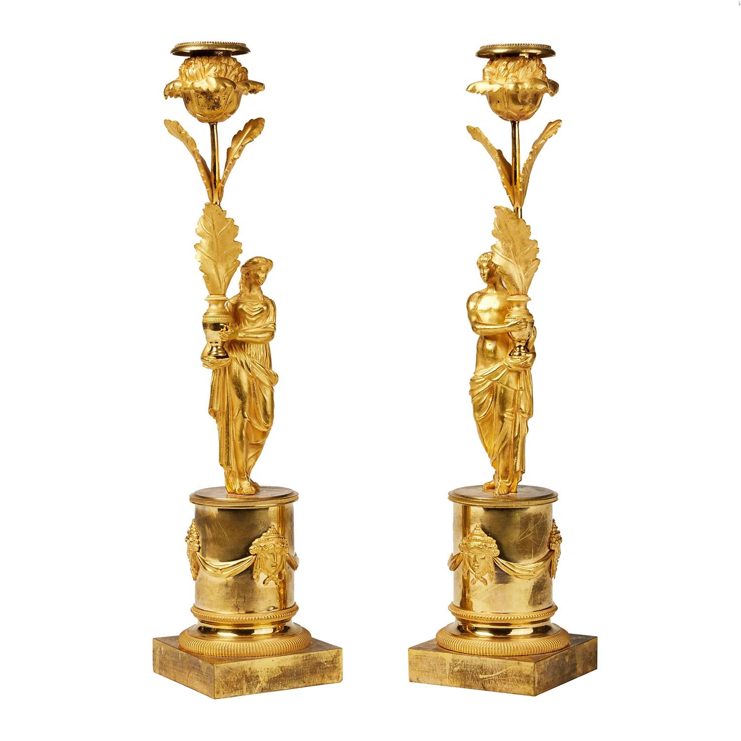 A gold, antique French pair of candle holders made of hand crafted matted and polished burnt gilded bronze, in good condition. Each of the detailed sticks is composed with a face mascarons, a crowned man and woman, carrying a mug with flowers, loose