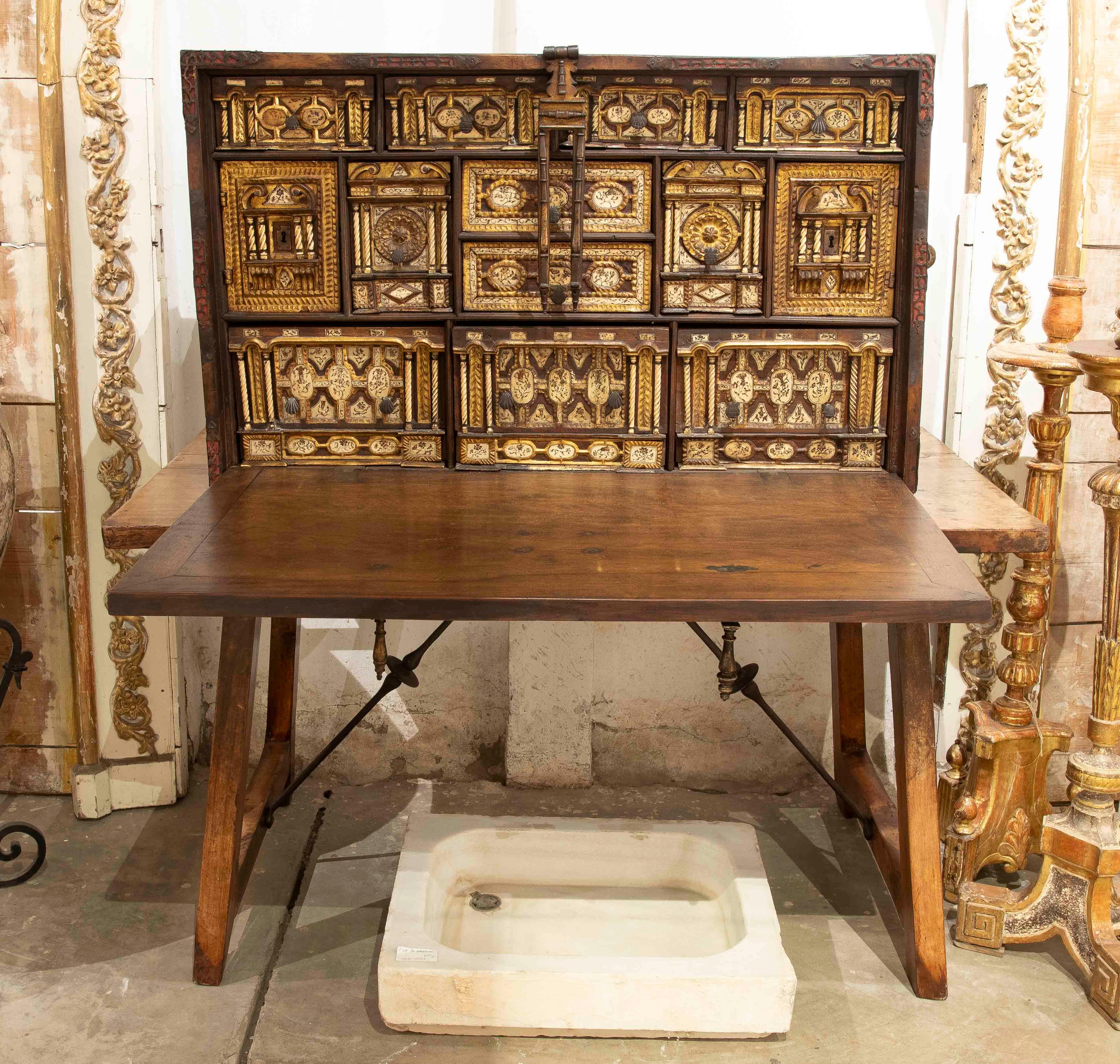 A majestic antique writing desk, richly carved and decorated with gilded details, placed on a sturdy, rustic wooden trestle-style table. The writing desk features multiple compartments and drawers, displaying a careful ornamentation of architectural
