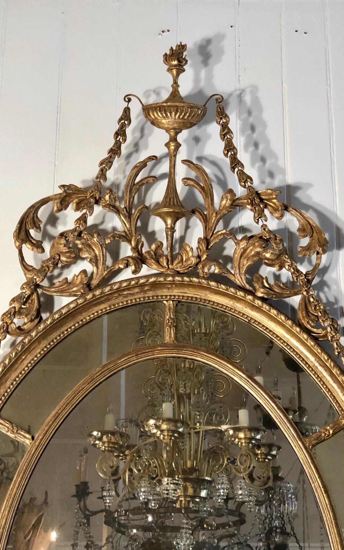 This elegant Adam mirror has a classical oval patera mirror border encompassing the looking glass. The crest has a central Adam urn with a burning flame flanked by scrolled leaves and branches coming up to a central reef below the urn. Chains of