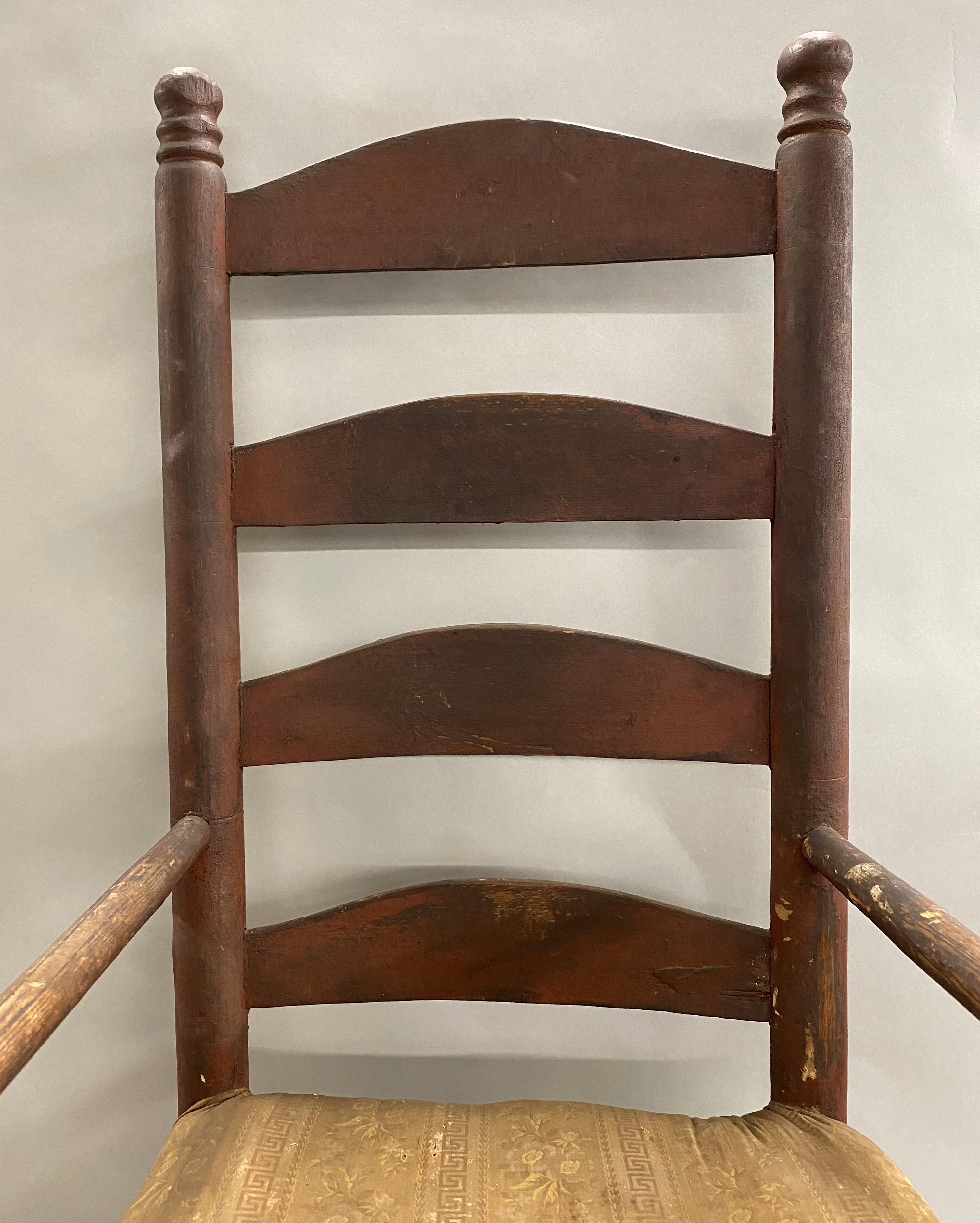 A wonderful old ladder back armchair in its original old crusty red paint, with great overall patina, nice wear to the arms, hand carved finials, and nice old faded cloth material on the padded seat. American in origin, dating to the 18th century,