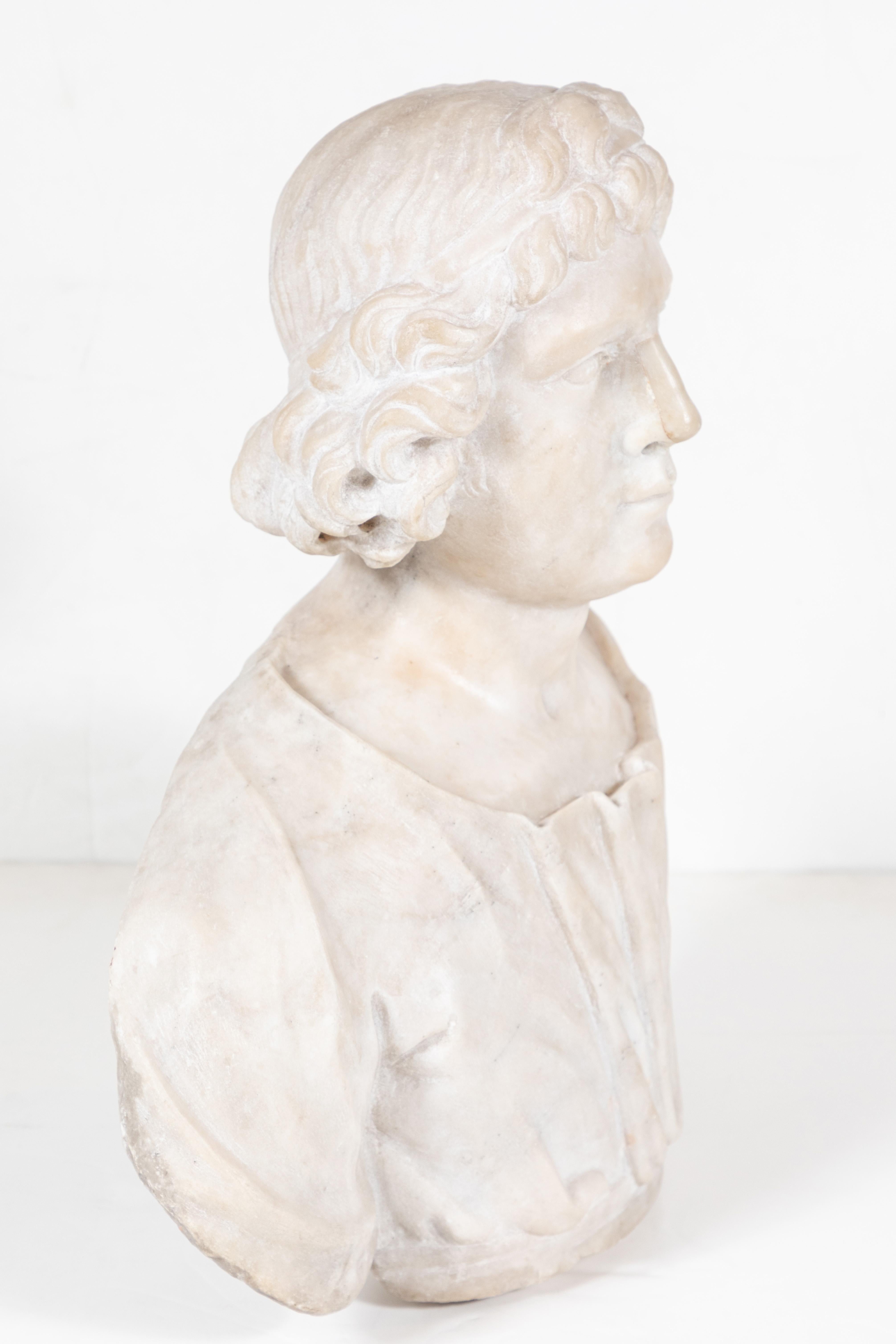 Hand carved, marble bust in three quarters, of a young man in a sensitively rendered, open-neck robe, with flowing hair held in a delicate circlet.