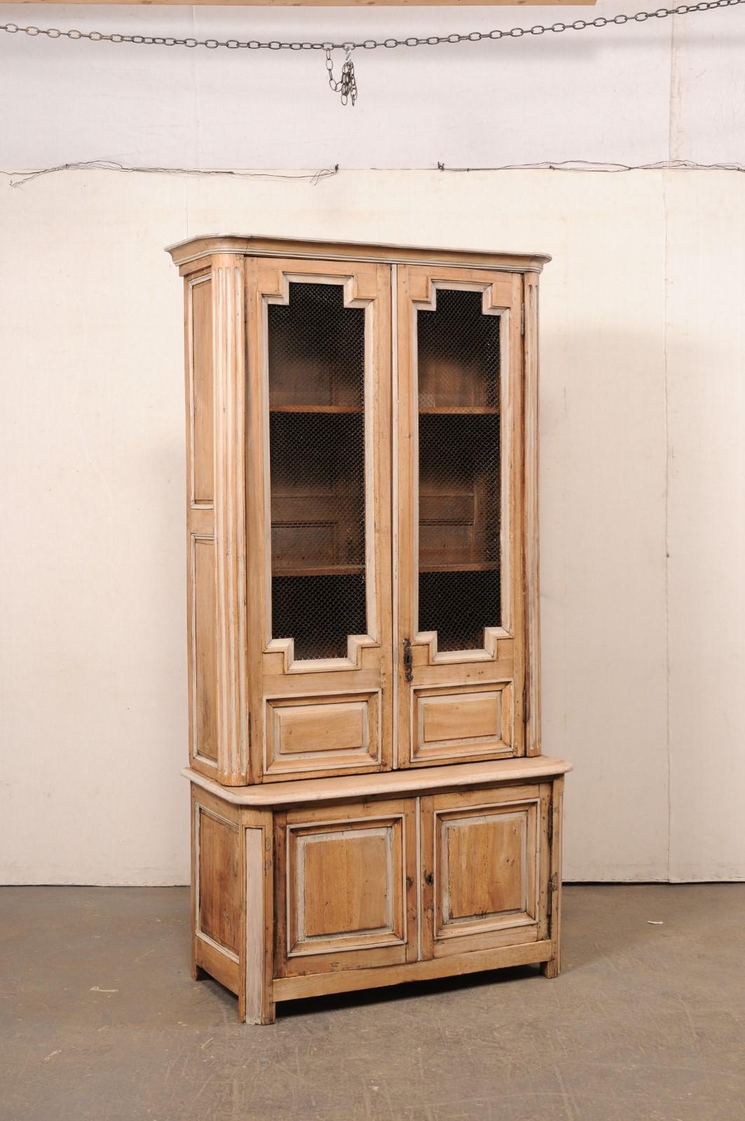 A tall French carved and bleached wood display and storage cabinet from the 18th century. This antique hutch cabinet from France features a beautifully geometric design motif with molded top crest over an upper case which houses a pair of wire-front