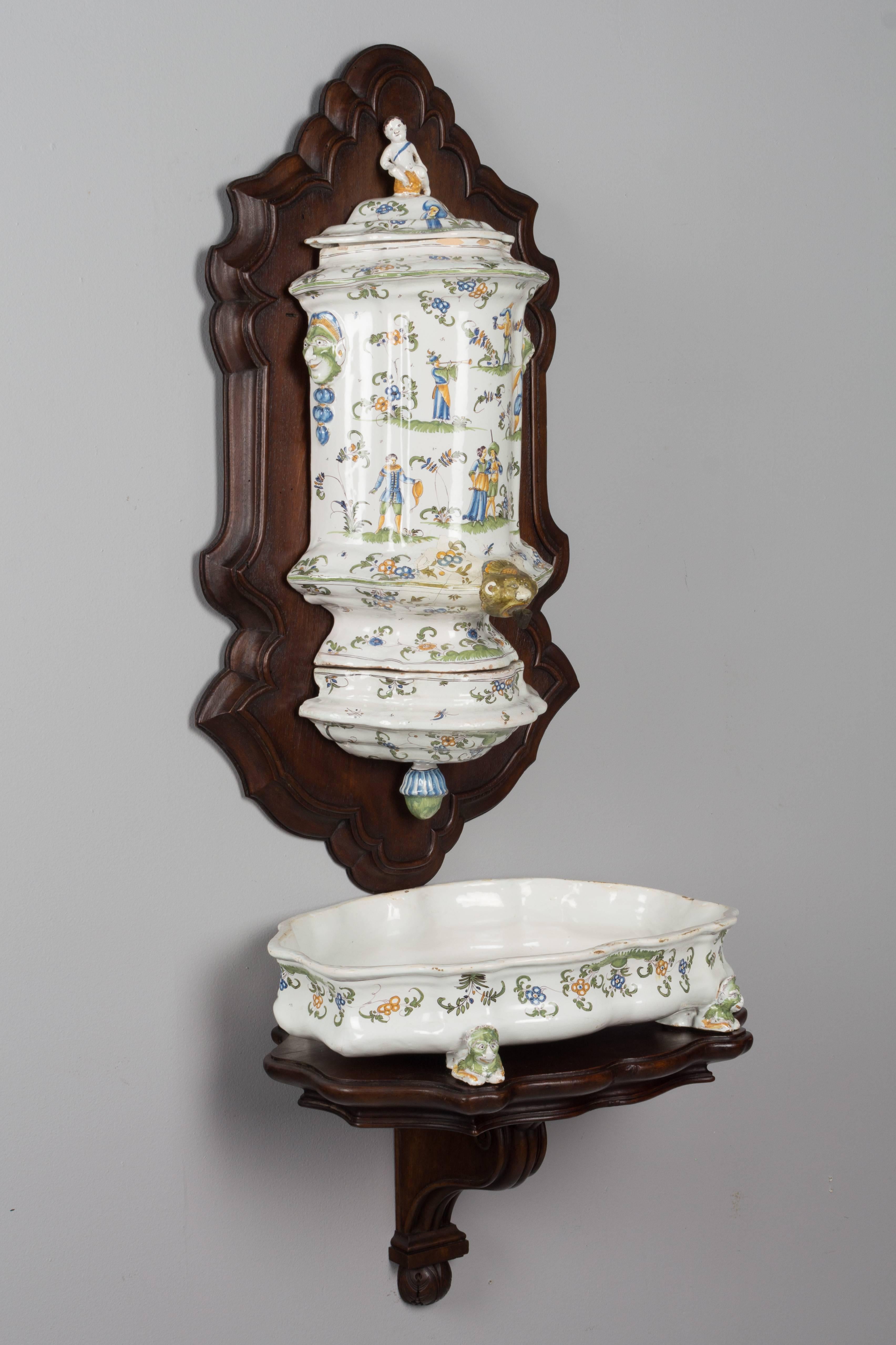 A French faience Louis XV style lavabo from Moustiers. The urn is in three parts that attach to a walnut plaque and the shallow basin rests on a walnut wall bracket. Hand-painted in blue, green and yellow with figures in a pastoral setting with