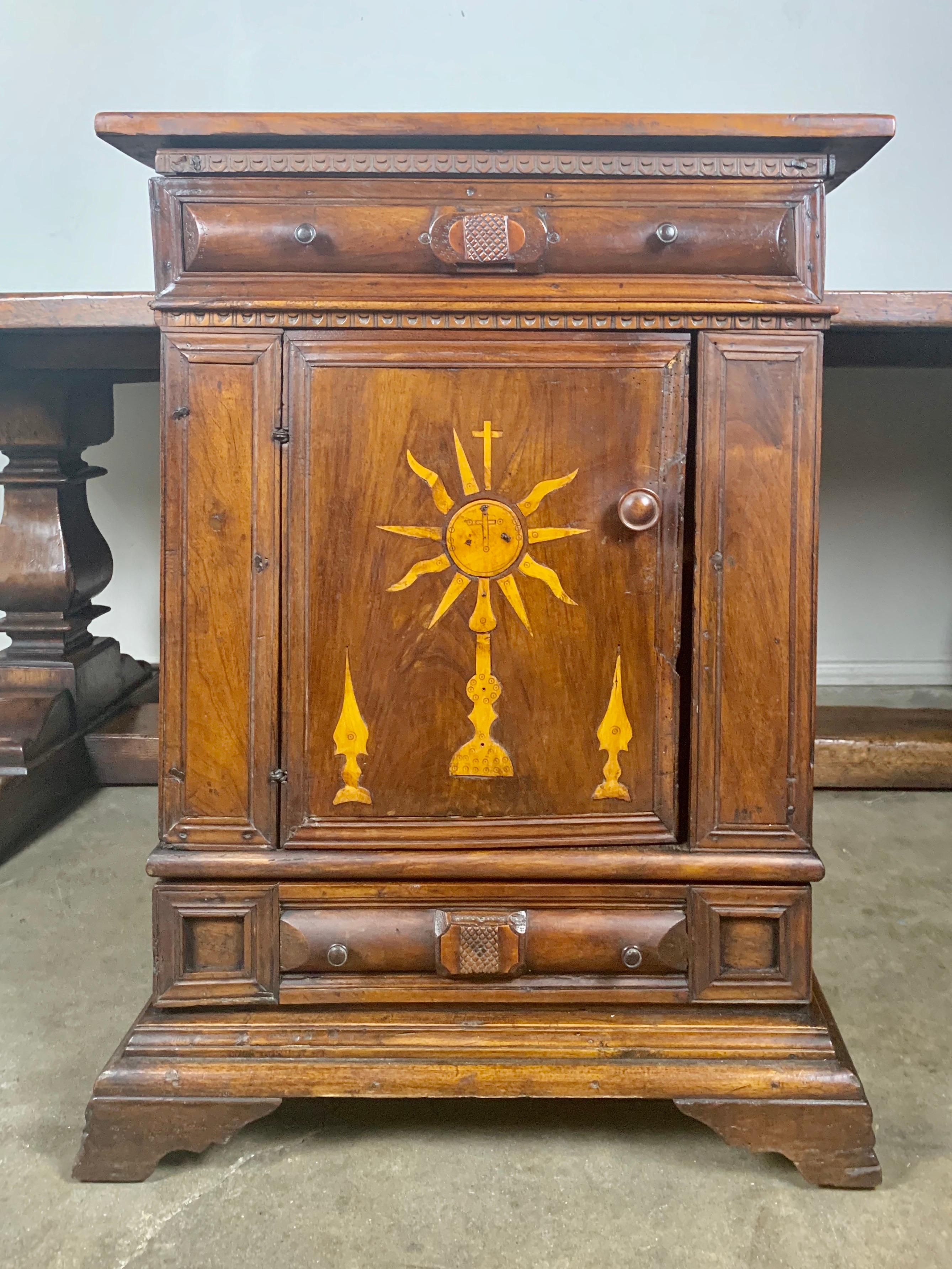 18th century inlaid Italian walnut credenza with maple inlay depicting various religious icons. There are two drawers, one towards the top and one on the bottom. There is a door that opens to more storage behind.