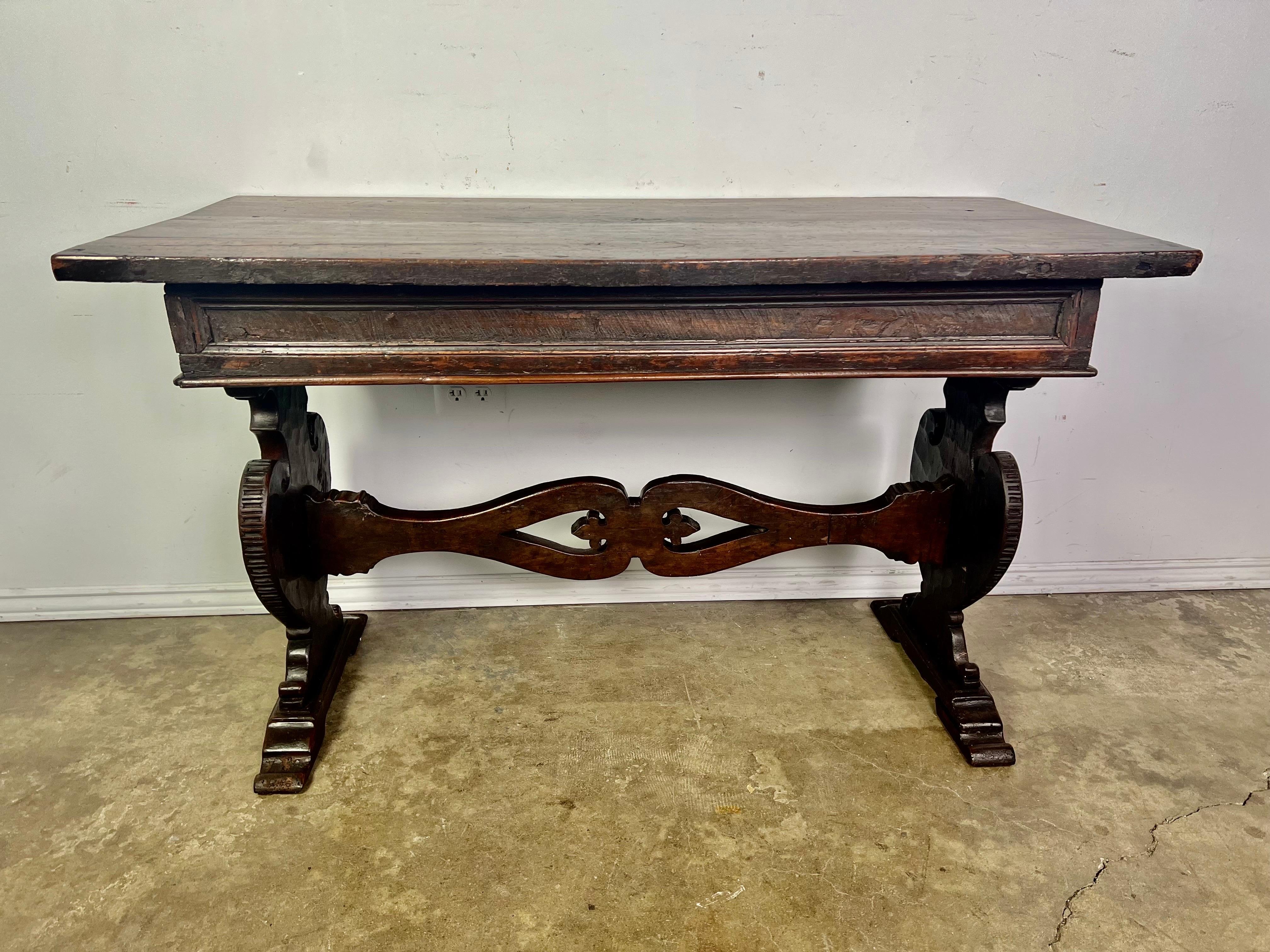 18th Century Italian walnut trestle style table. A rectangular top sits on a pair of lyre shaped bases connected by a center stretcher. The stretcher is hand carved with fleur dy lis within a pair of connecting loops. The table has developed a