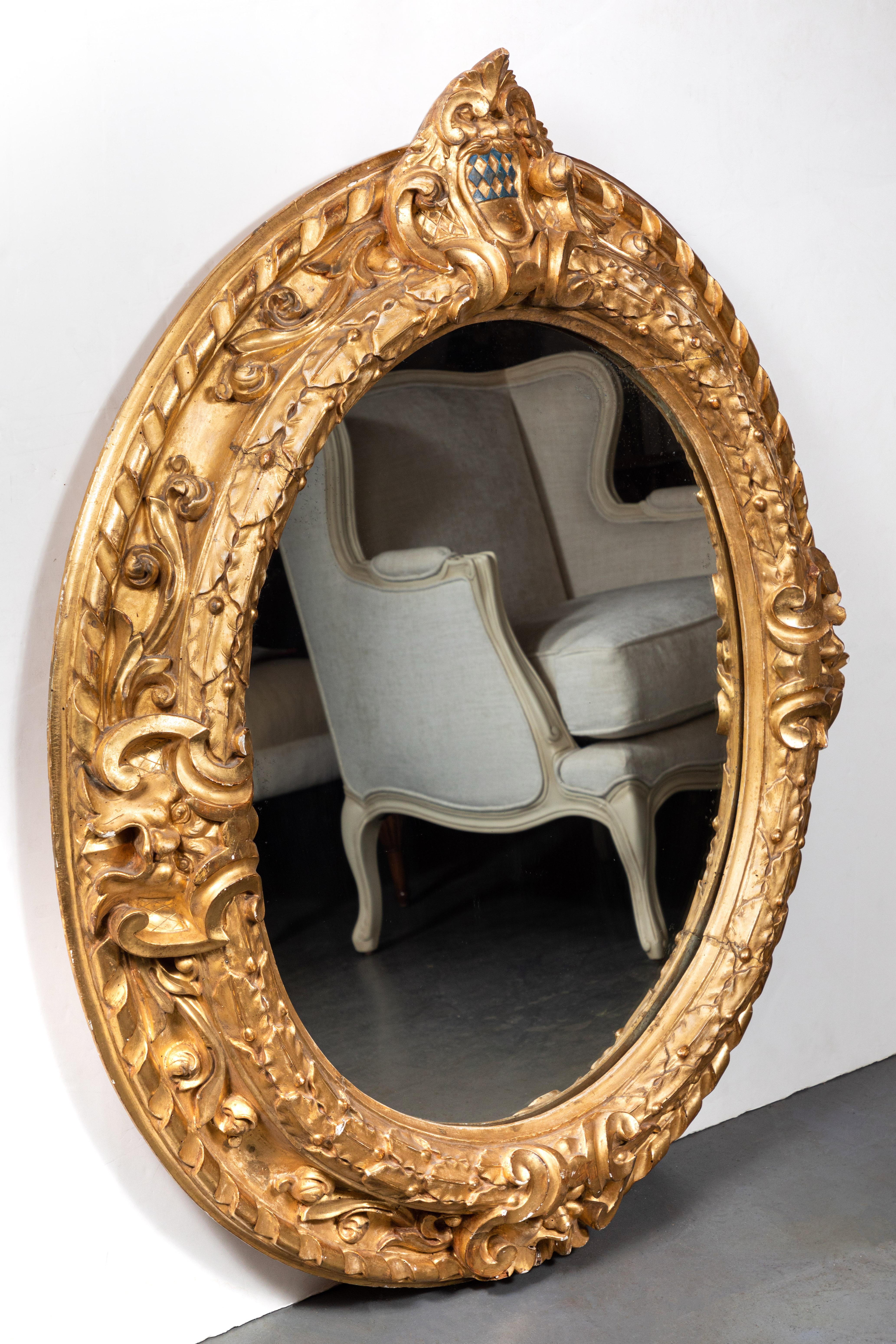 Grand cadre ovale italien, sculpté à la main et doré à l'or fin 22 carats, avec miroir encastré plus récent. La couronne présente un écusson peint à la main avec un motif matelassé de diamants verts. L'ensemble est orné de formes feuillues.