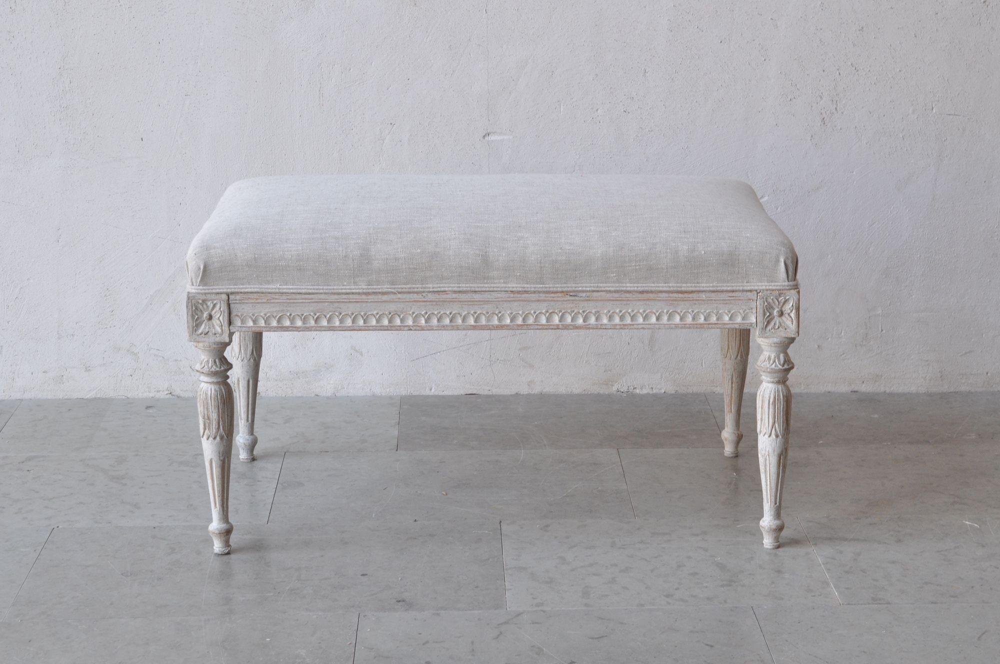 This is a richly carved 18th Century Swedish Gustavian period painted bench, newly upholstered in linen. There is carved egg and dart detail on the seat frame and carved rosettes above round, tapered and fluted legs adorned with acanthus leaves.
