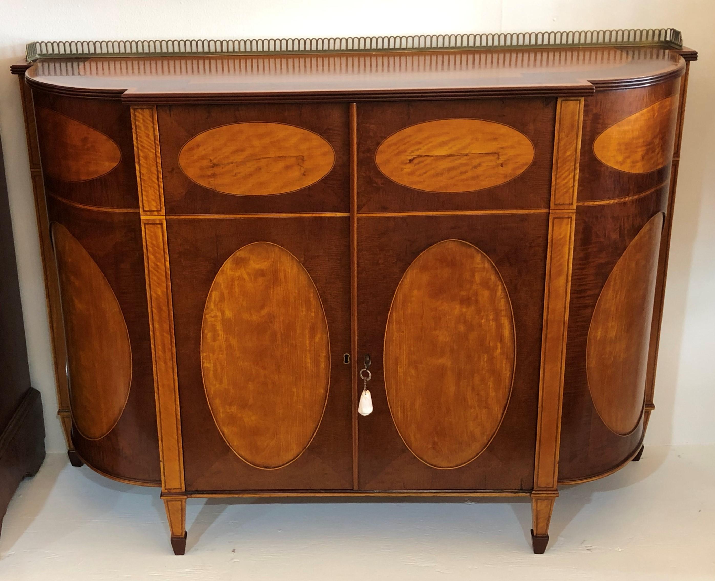 This elegant satinwood commode is inlaid with purple wood, mahogany, and ebony has two doors with satinwood ovals. The neoclassical credenzas legs are satinwood with ebony string inlay finishing into applied mahogany spade feet. The top has a