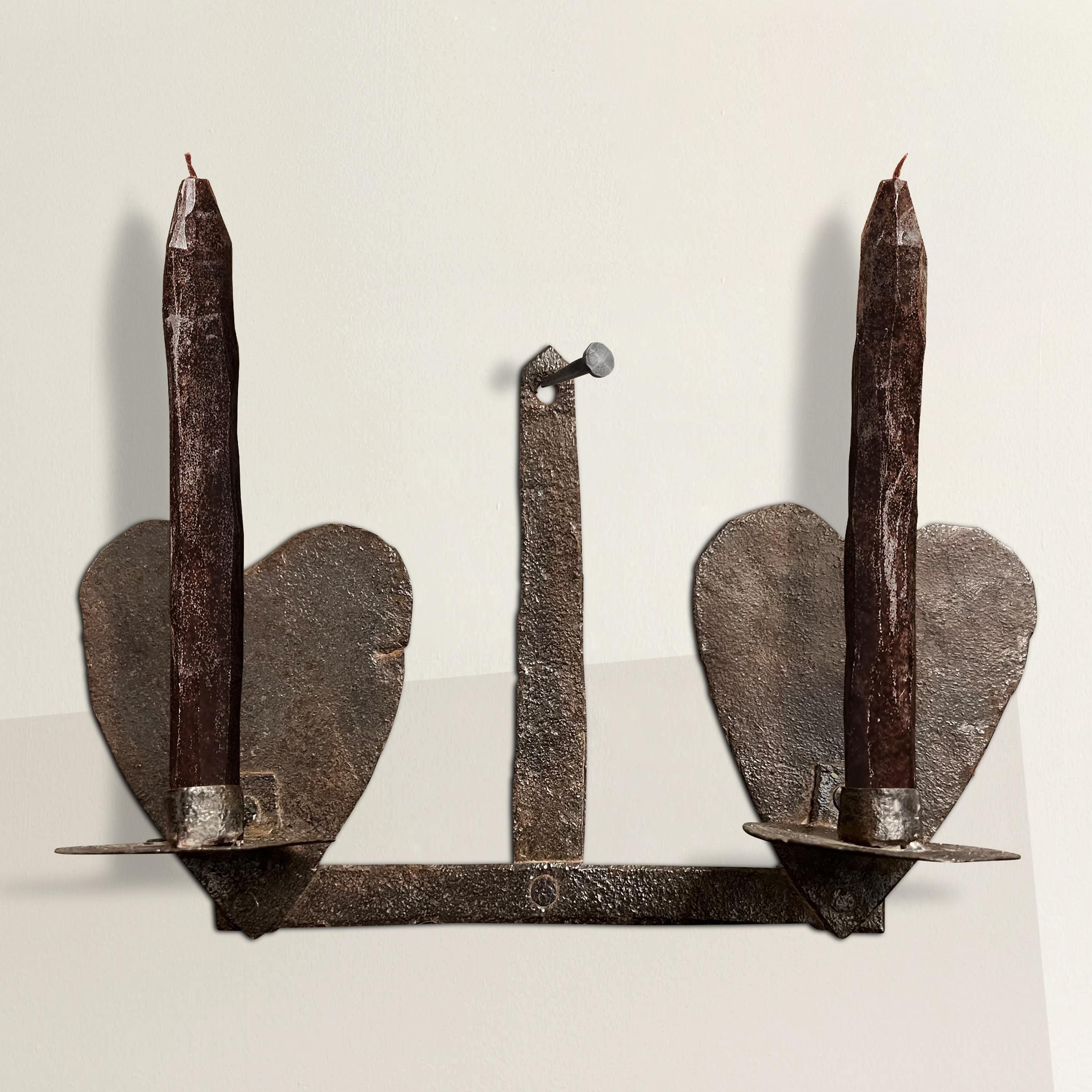 Transport yourself to 18th century America with this primitive wrought iron two-arm candle sconce, a testament to both functionality and aesthetic simplicity. The heart-shaped backplates exude a charming rustic elegance, capturing the essence of