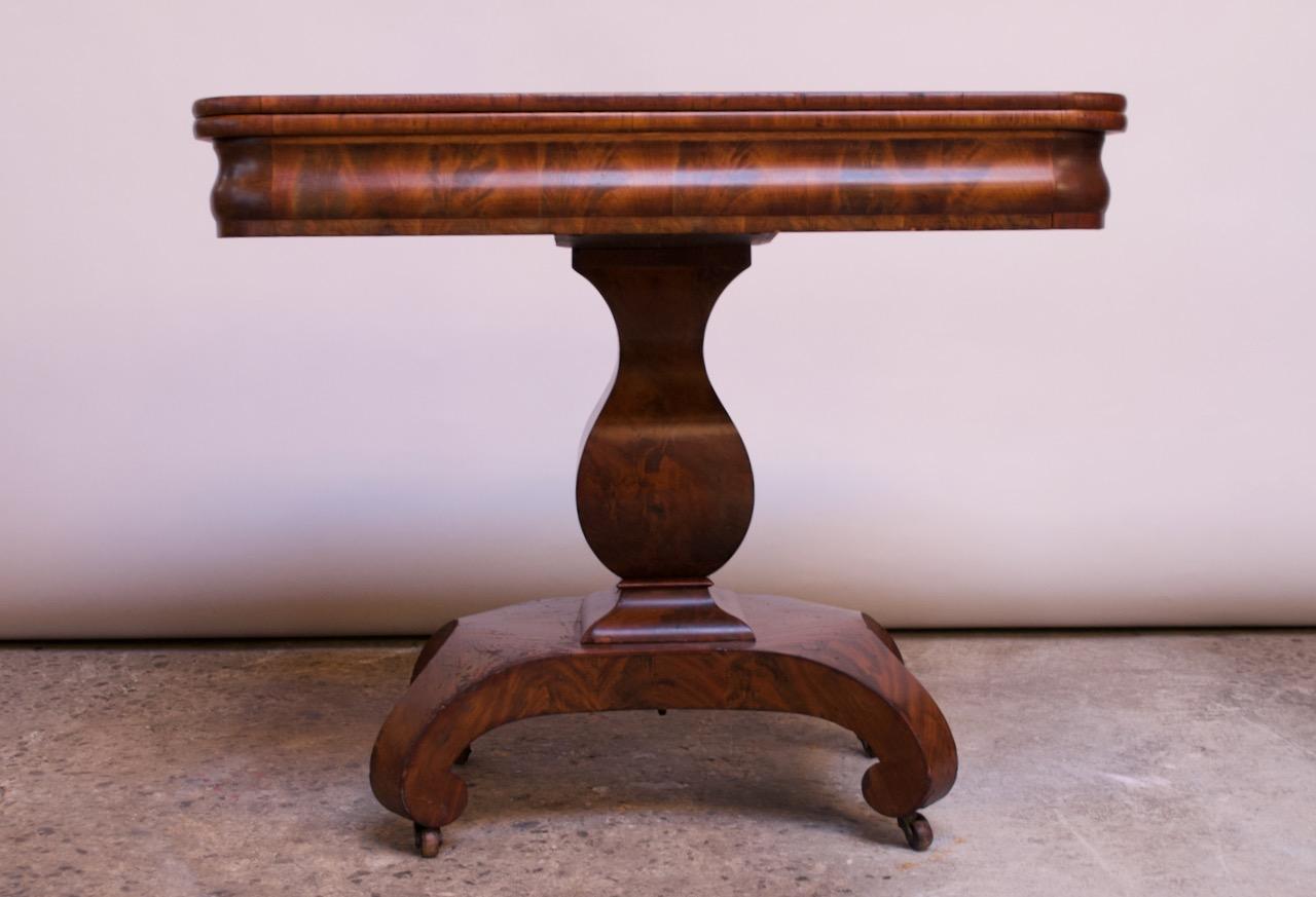 American Empire matchbook flame mahogany game or parlor table, circa mid-1700s. Features a hinged top concealing a hidden compartment which flips over to create a larger, square surface. Top is supported by a pedestal base with scroll feet and