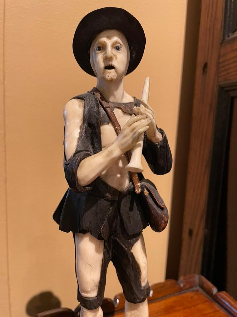 Carved bone and walnut figure of a beggar with a very expressive face holding a flute, with a satchel over his shoulder carved from wood with a leather strap. With inset glass eyes. On an integrally carved walnut base. 
Workshop of Simon Troger