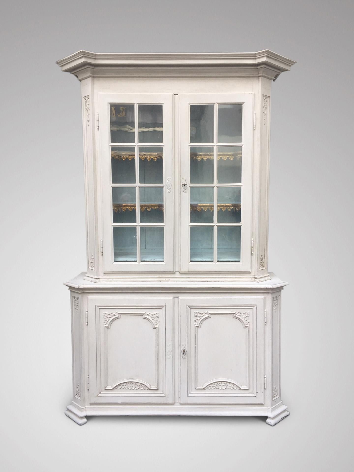A mid-18th century, Belgian from the Liège town, white painted oak display cabinet. Carved overall with C-scrolled rock-work and flower baskets, the eared moulded breakfront cornice above a pair of original glazed doors enclosing a light blue