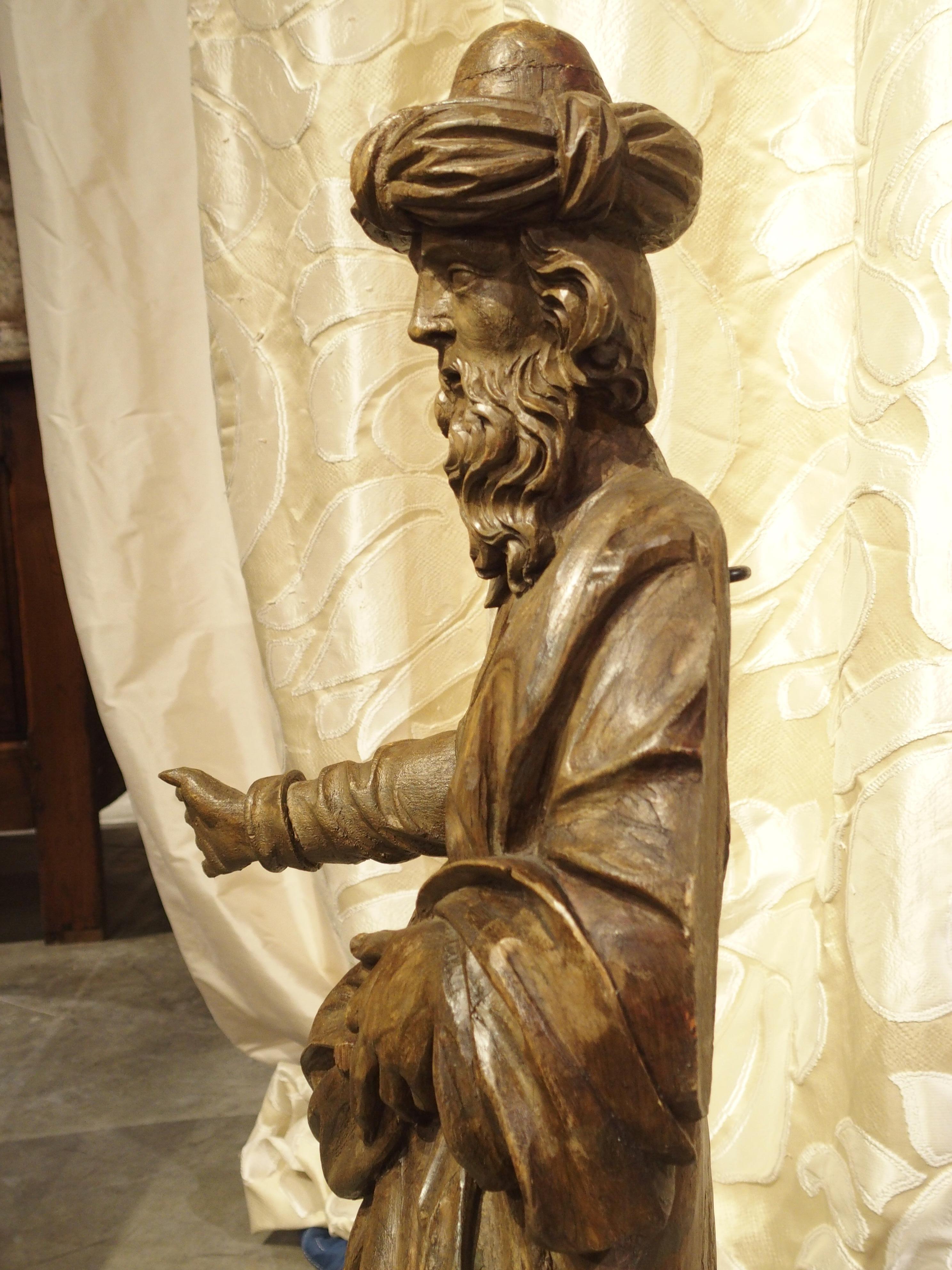This antique French hand carved wooden statue is of a bearded man wearing a robe tied at the waist with a long scarf draped over his shoulder. The back of the statue has an opening where it was likely attached to something else at one point in time.