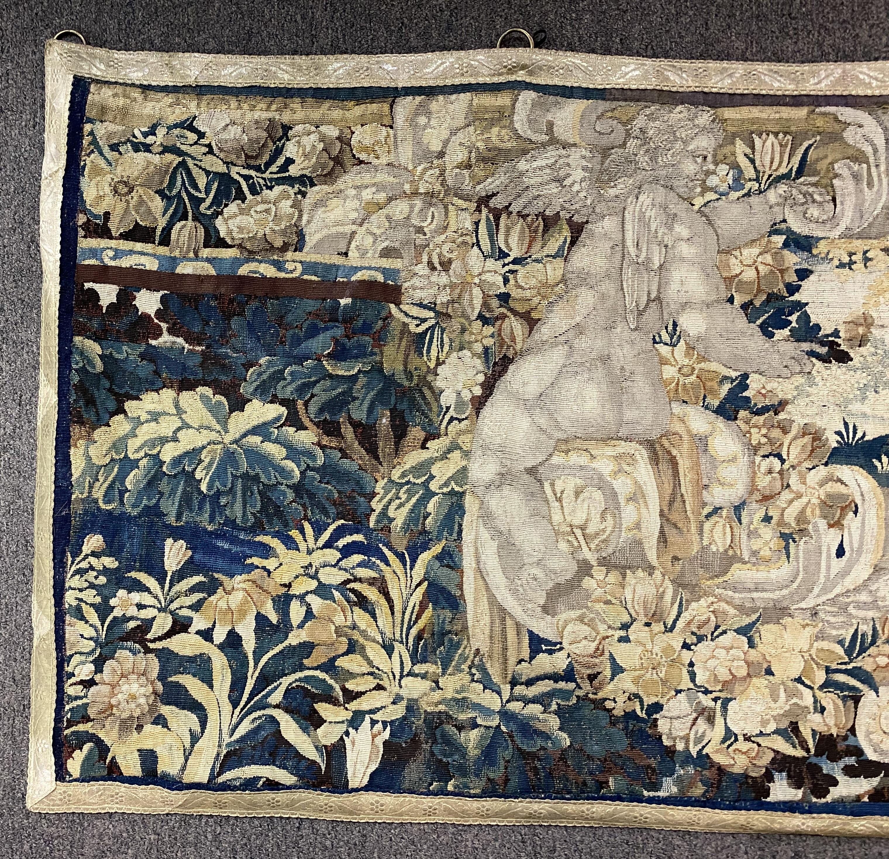 A nice example of a Continental hand wrought tapestry decorated with putti or angels and cartouche, with foliate accents and subtle tan border with raised flowers. The tapestry has a plain fabric backing, and brass rings have been added along the