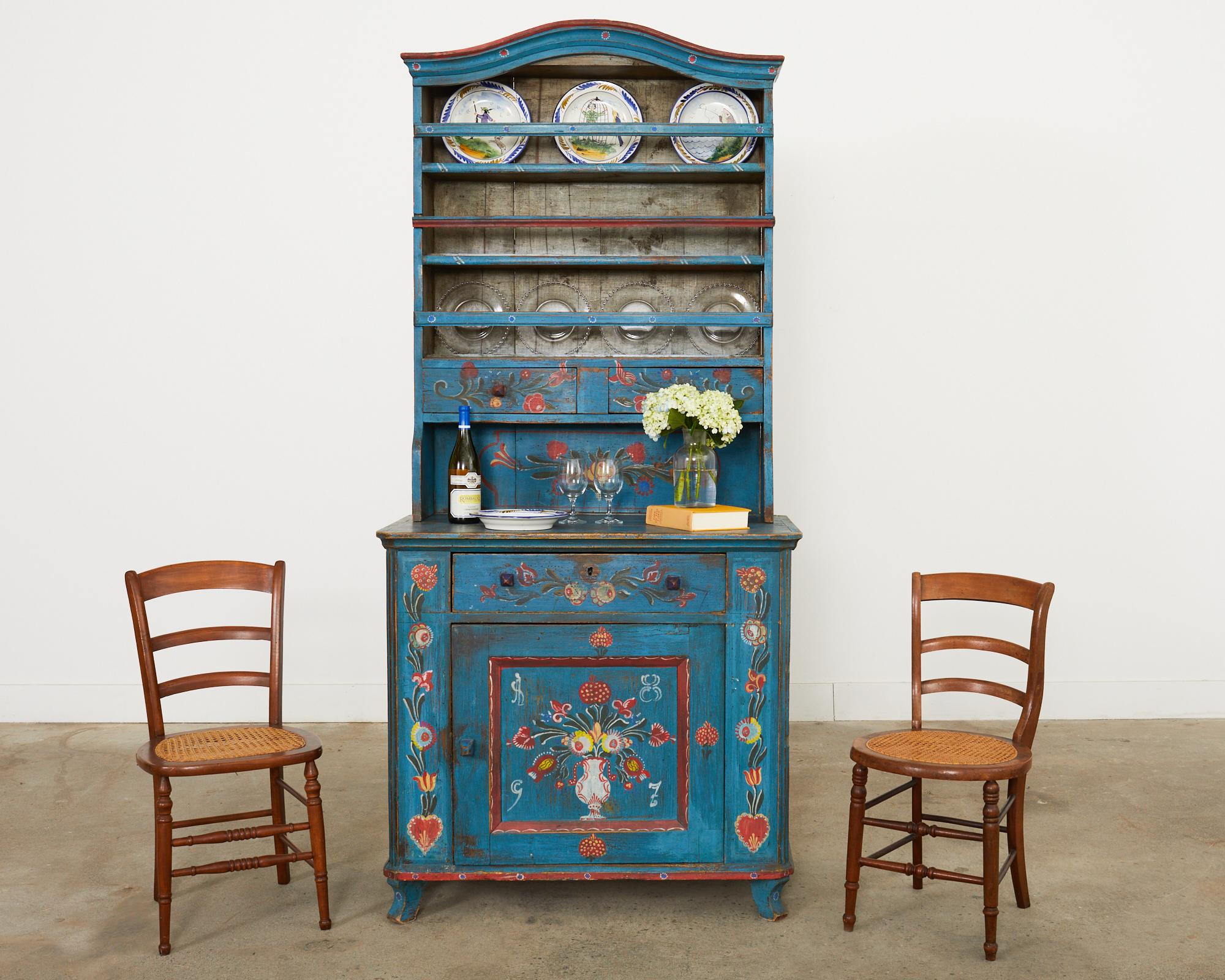 Whimsical 19th century country Swedish painted farmhouse cabinet dresser topped with a cupboard plate rack. The cabinet features a colorful festive folk art style painted finish with a floral motif centered by a vase with a floral spray. Crafted