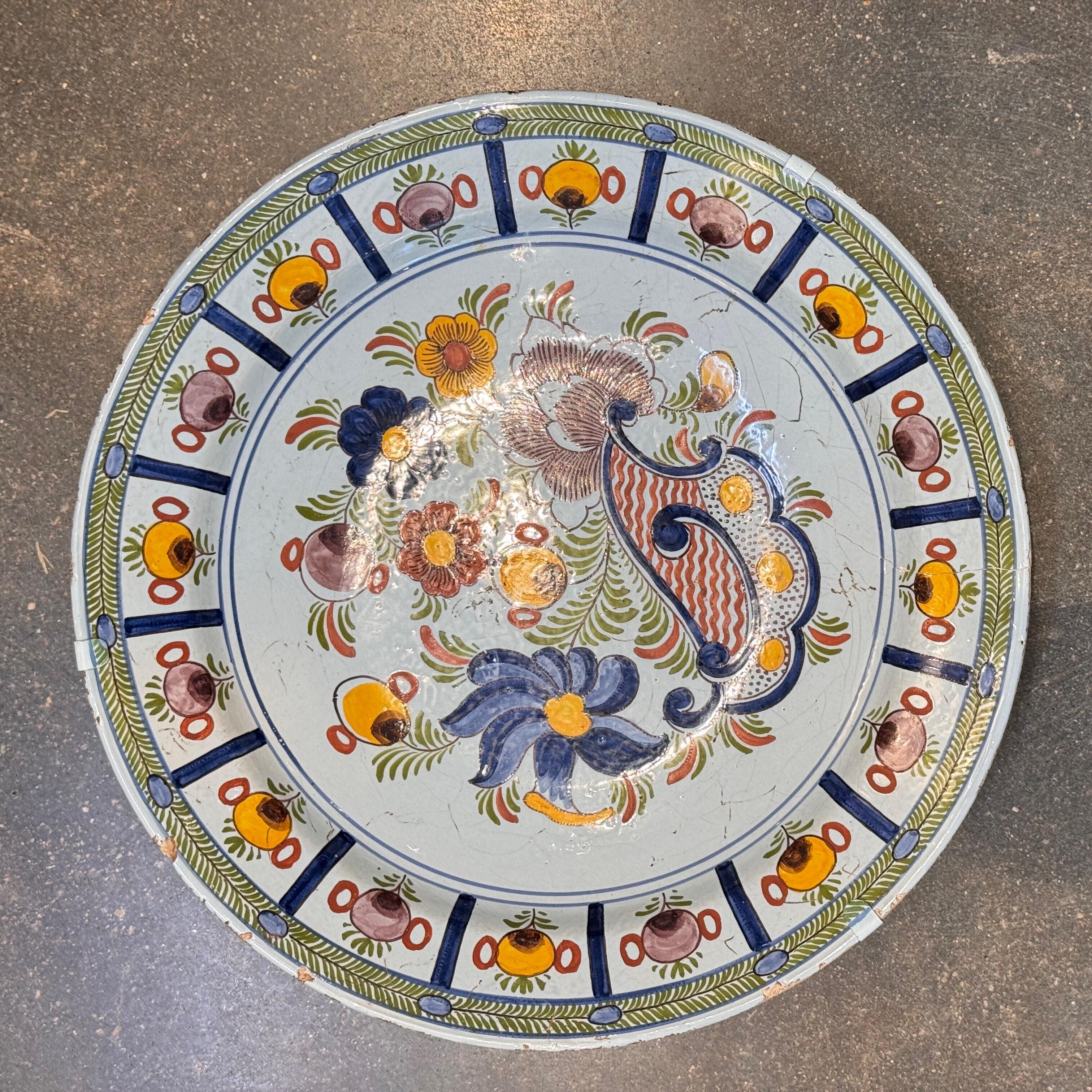 Gran jarrón de Delft del siglo XVIII pintado a mano en vibrante policromía, con un ramo central de flores estilizadas y un borde festoneado de urnas y follaje. La superficie de estaño vidriado presenta grietas propias de la edad, y el plato lleva un