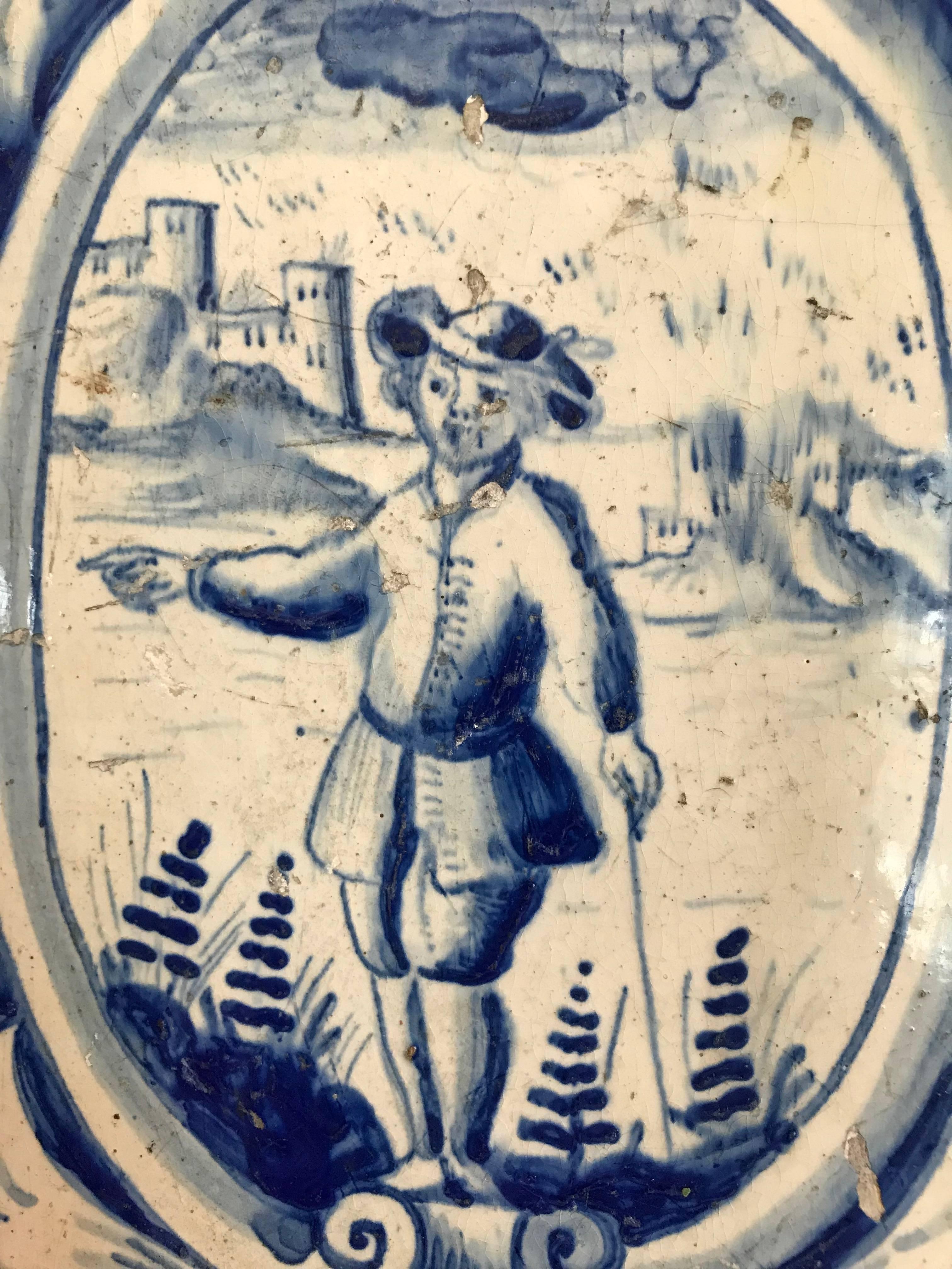 Large-scale Dutch delft stove tile with a portrait of a man with a walking stick wearing typical 18th century costume, behind him a landscape with castle. All within a cartouche framed with floral scroll work above and below.
Mounted on a removable