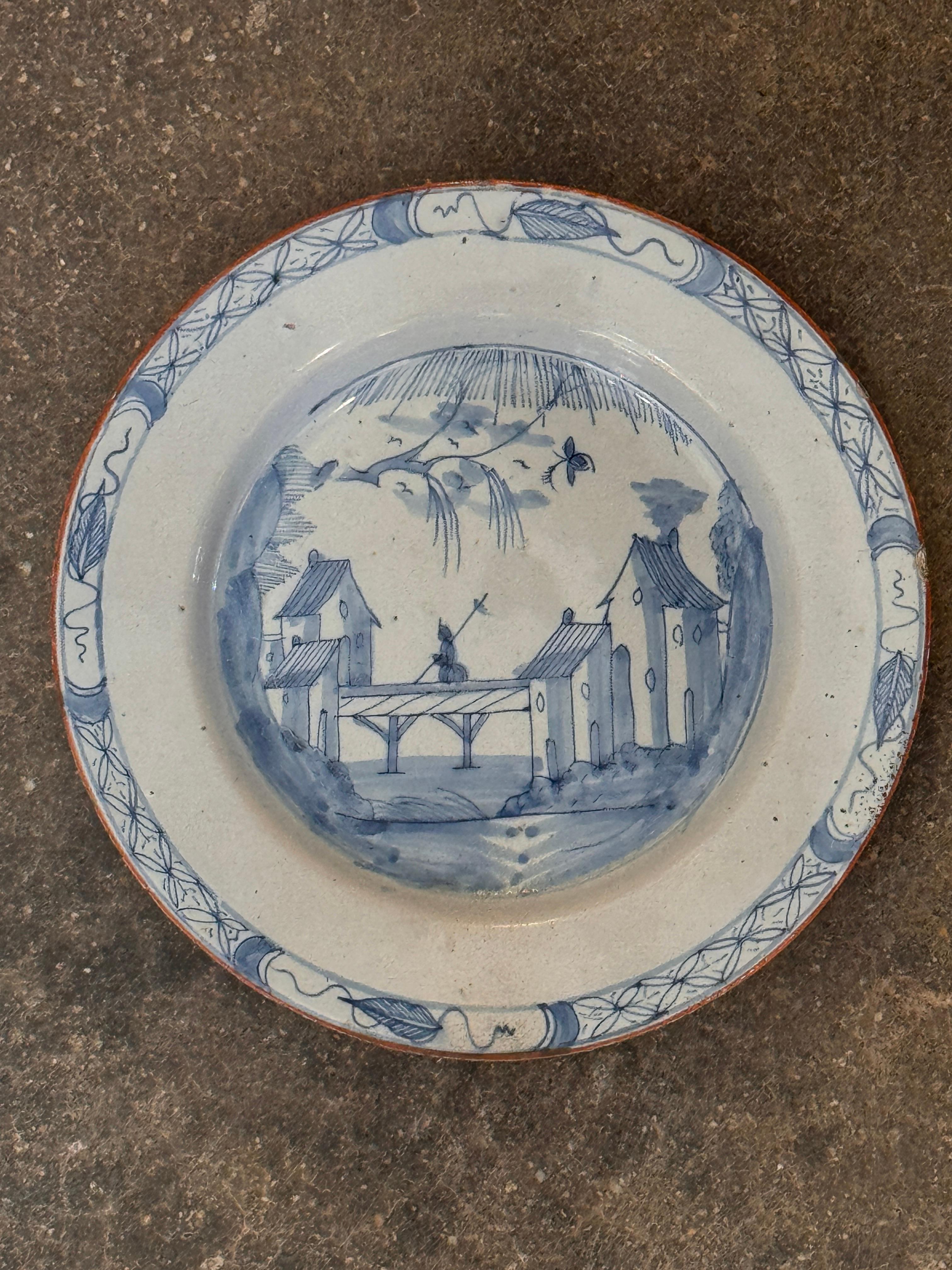 Piatto di Delft del XVIII secolo dipinto a mano in blu e bianco con una scena centrale di edifici e un ponte sotto un salice, racchiusa da un bordo decorativo. La superficie smaltata a stagno presenta un'usura tipica delle prime ceramiche di Delft.
