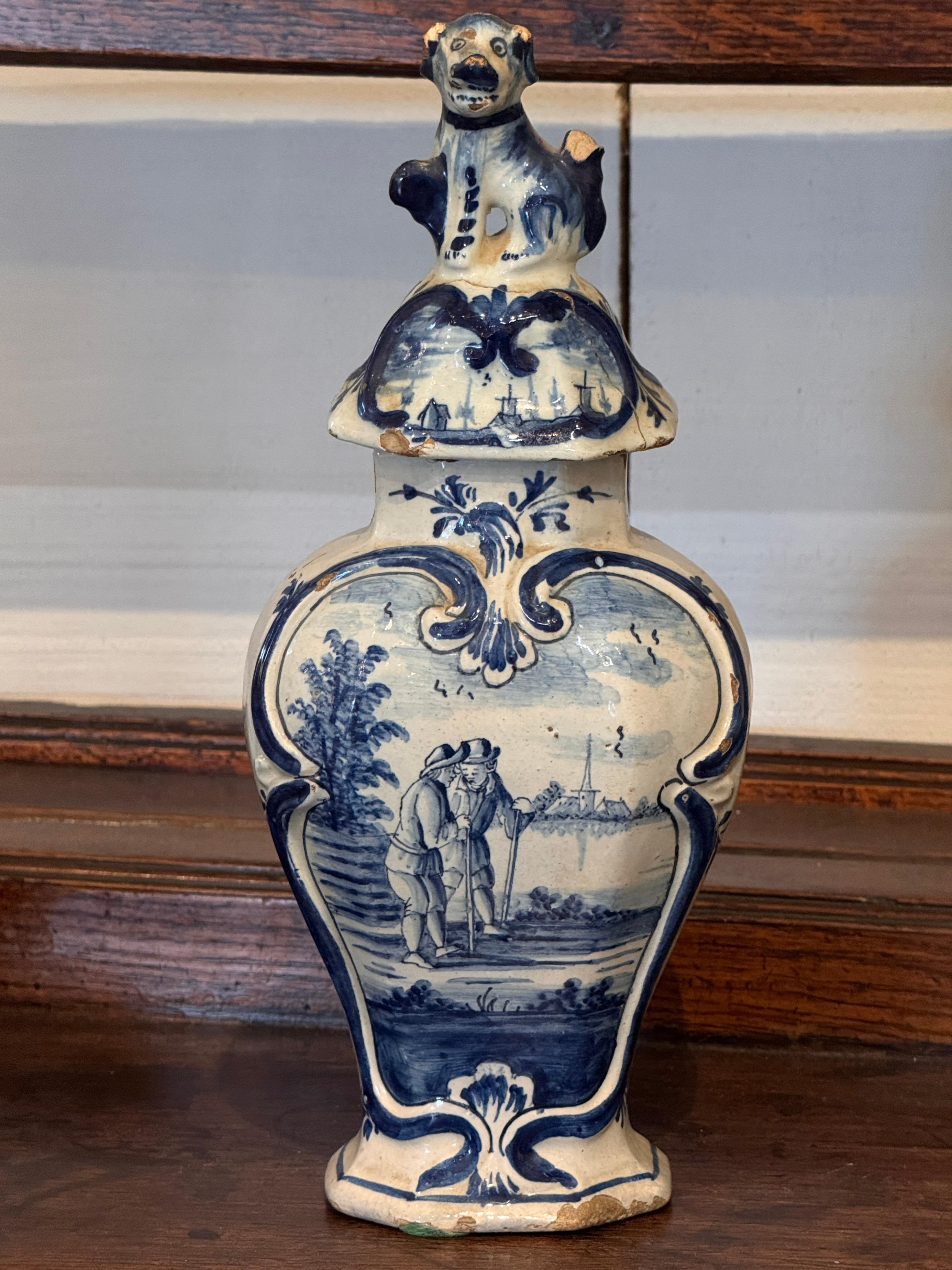 Vase à couvercle en faïence de Delft bleu et blanc, XVIIIe siècle, décoré de scènes de paysages pastoraux encadrées dans des cartouches. Le couvercle comporte un fleuron figuratif en forme d'animal assis, ajoutant un détail vivant. Les scènes