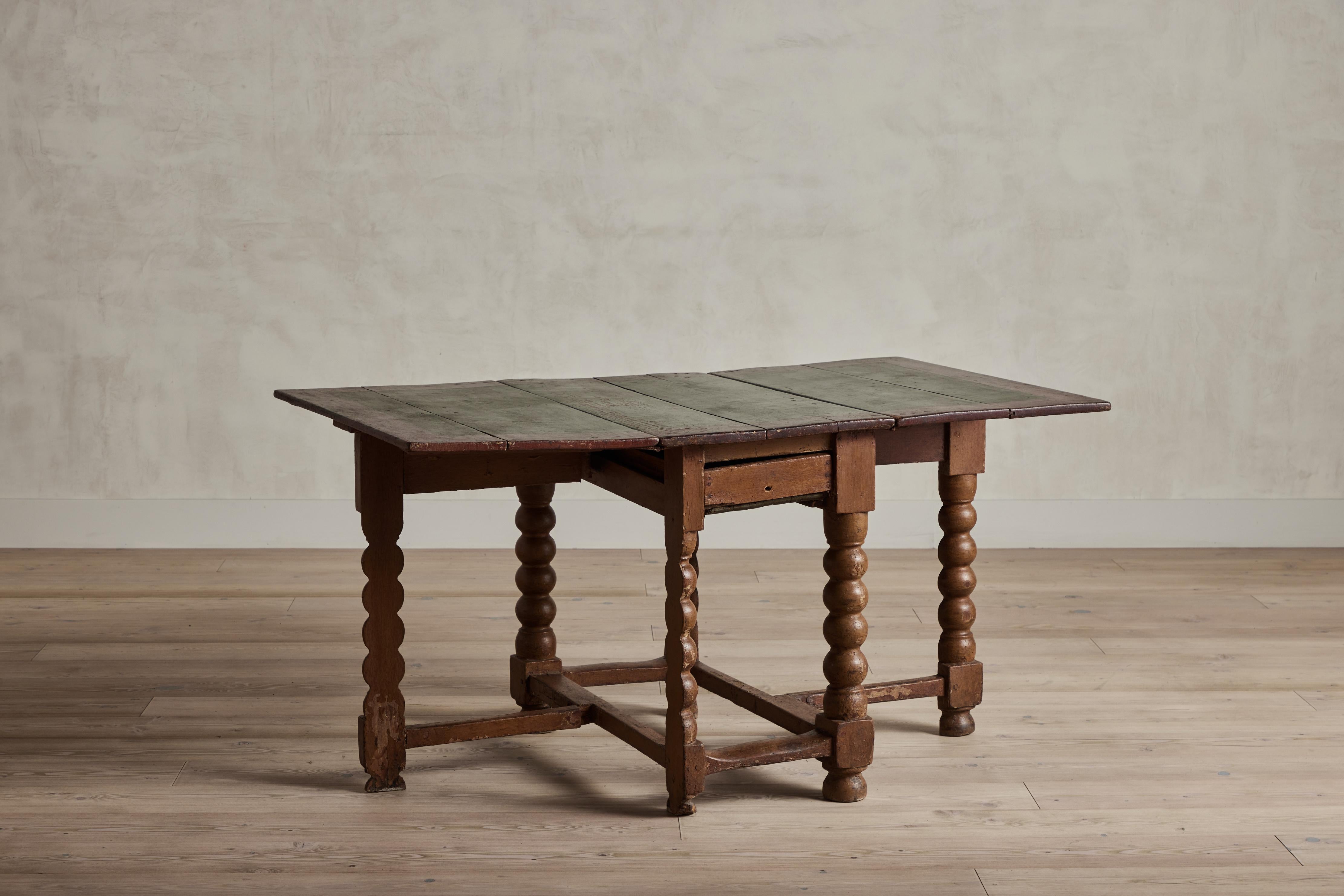 Table hollandaise du XVIIIe siècle en bois avec un plateau rustique peint en vert et rouge. La table possède un petit tiroir central qui se retire. Les pieds articulés s'ouvrent pour supporter les feuilles qui prolongent la surface de la table.