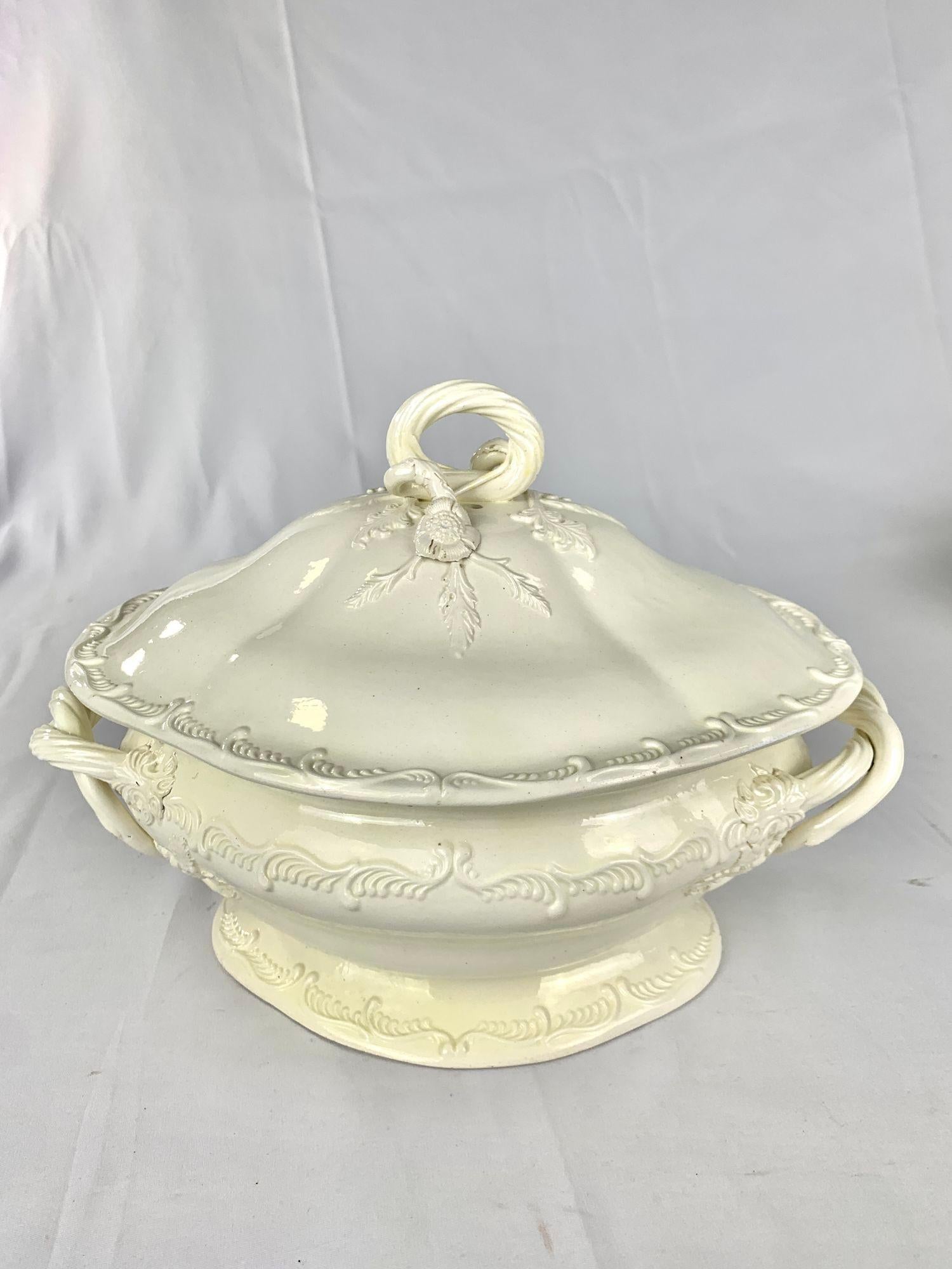 Esta sopera de cerámica cremosa, rara y bellamente modelada, fue fabricada por Leeds Pottery hacia 1780.
Presenta una tapa abovedada rematada con un remate de lazo adornado con flores y follaje aplicados.
El cuerpo está nítidamente moldeado con tres