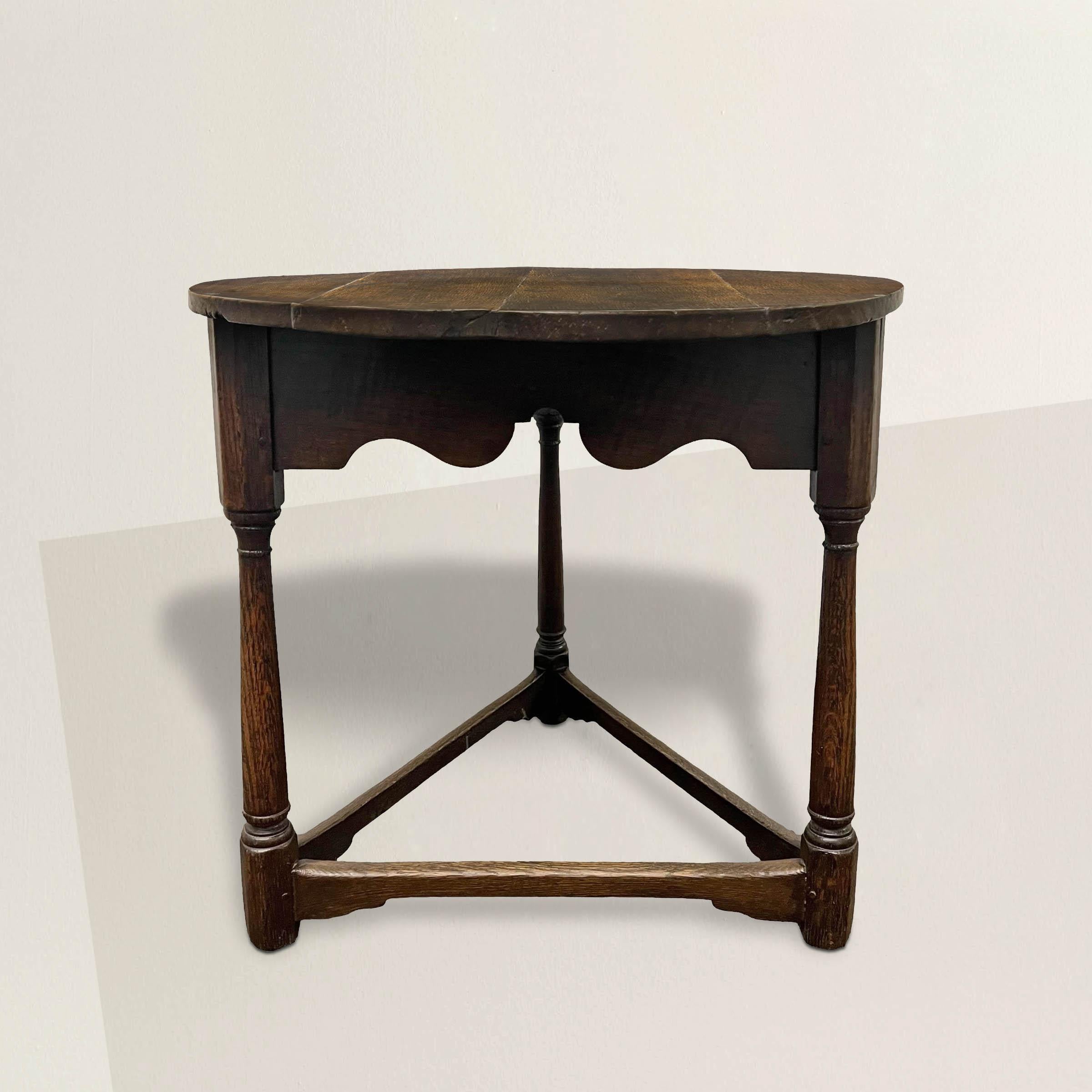 Behold the timeless elegance of this 18th-century English oak cricket table, a quintessential piece of English culture. Its round top, adorned with a scalloped apron, rests gracefully atop three exquisitely turned columnar legs, showcasing masterful