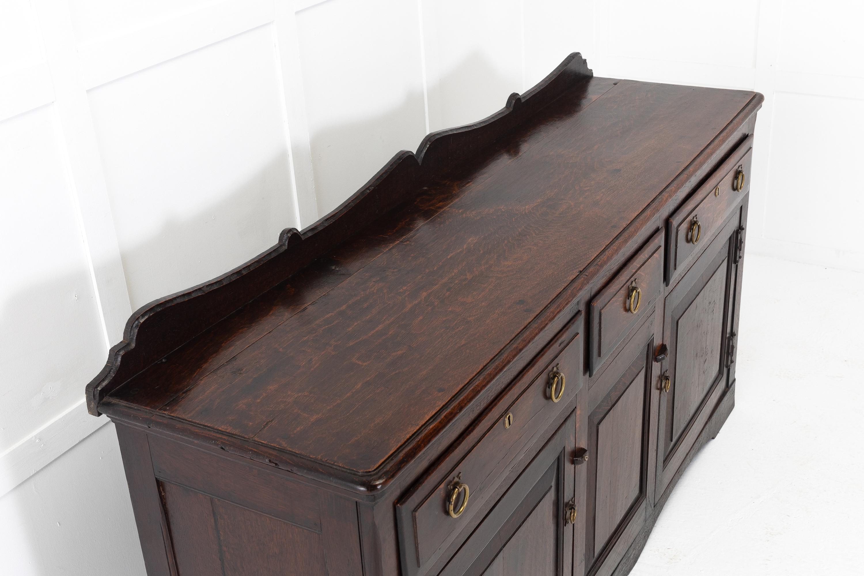 18th Century English George III oak dresser base with shaped gallery back. Having three drawers across the top with brass ring pull handles and escutcheons. The full cupboard below the drawers has panelled cupboard doors at each end to access the