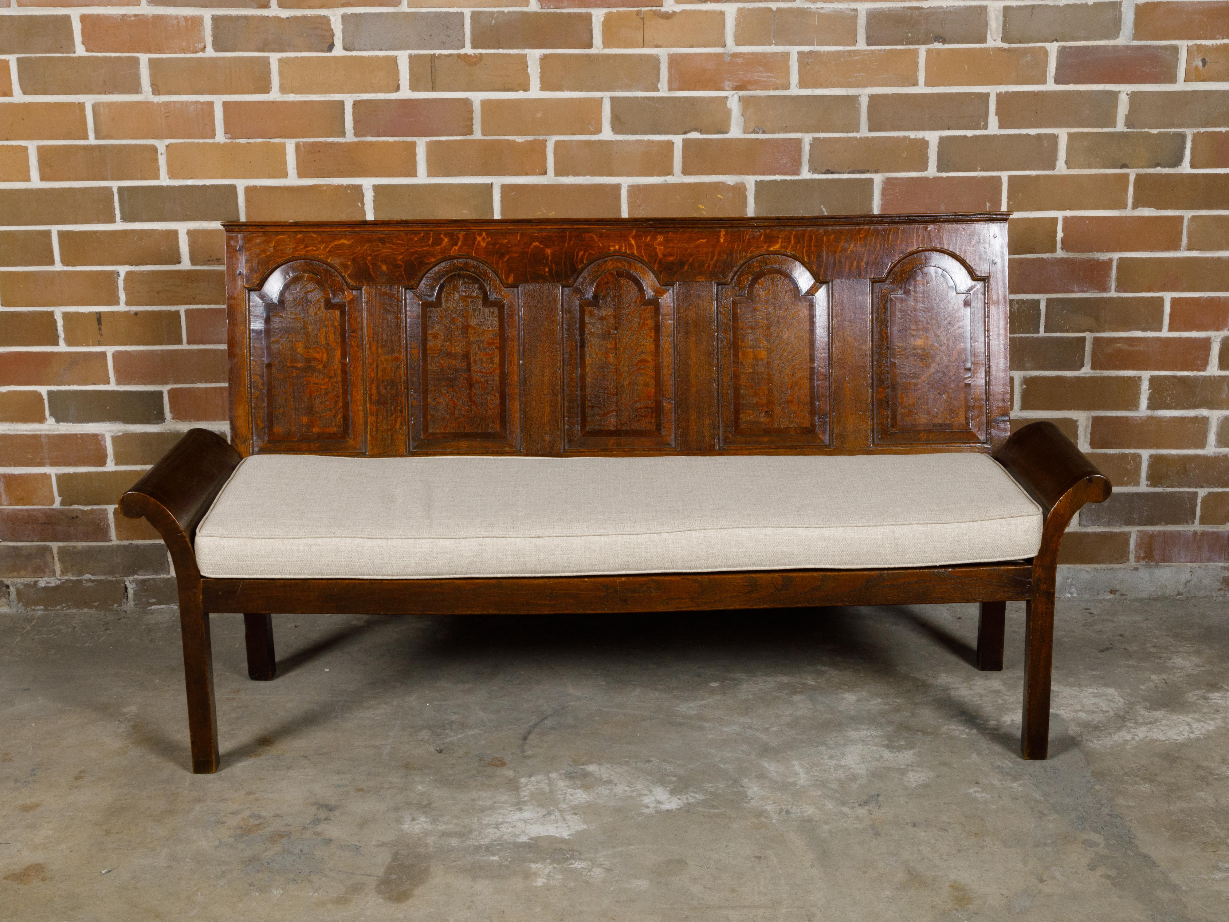 An English Georgian period oak bench from the 18th century with solid carved back, out-scrolling arms and new custom upholstered cushion. This exquisite English Georgian period oak bench from the 18th century is a remarkable piece of furniture that