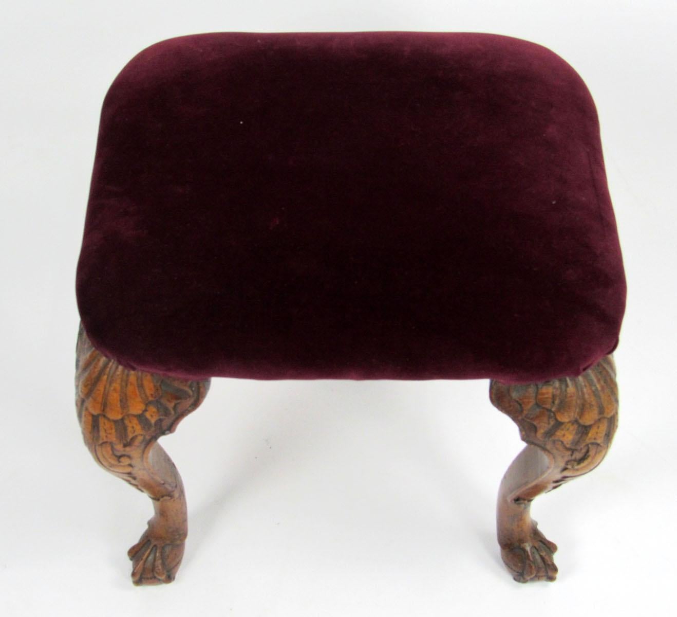 18th century English mahogany footstool with a shell and scroll design carved into the legs above ball and claw feet, and upholstered in a burgundy velvet fabric.