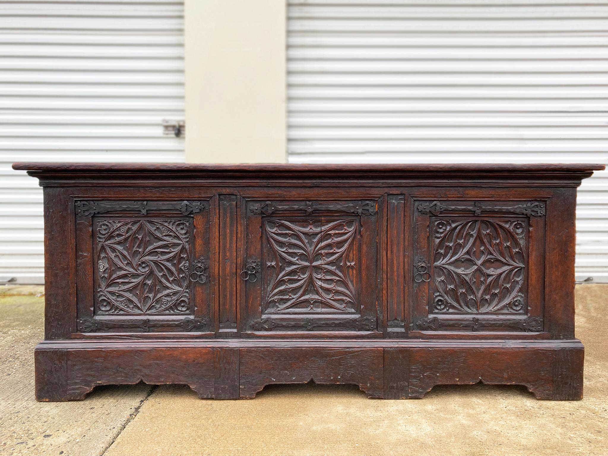 18th century English coffer trunk, handcrafted from oak. Front panels feature ornate, hand-carved floral and geometric designs. The wood has a warm red tone and has aged beautifully. The iron hinges of the lid are a later