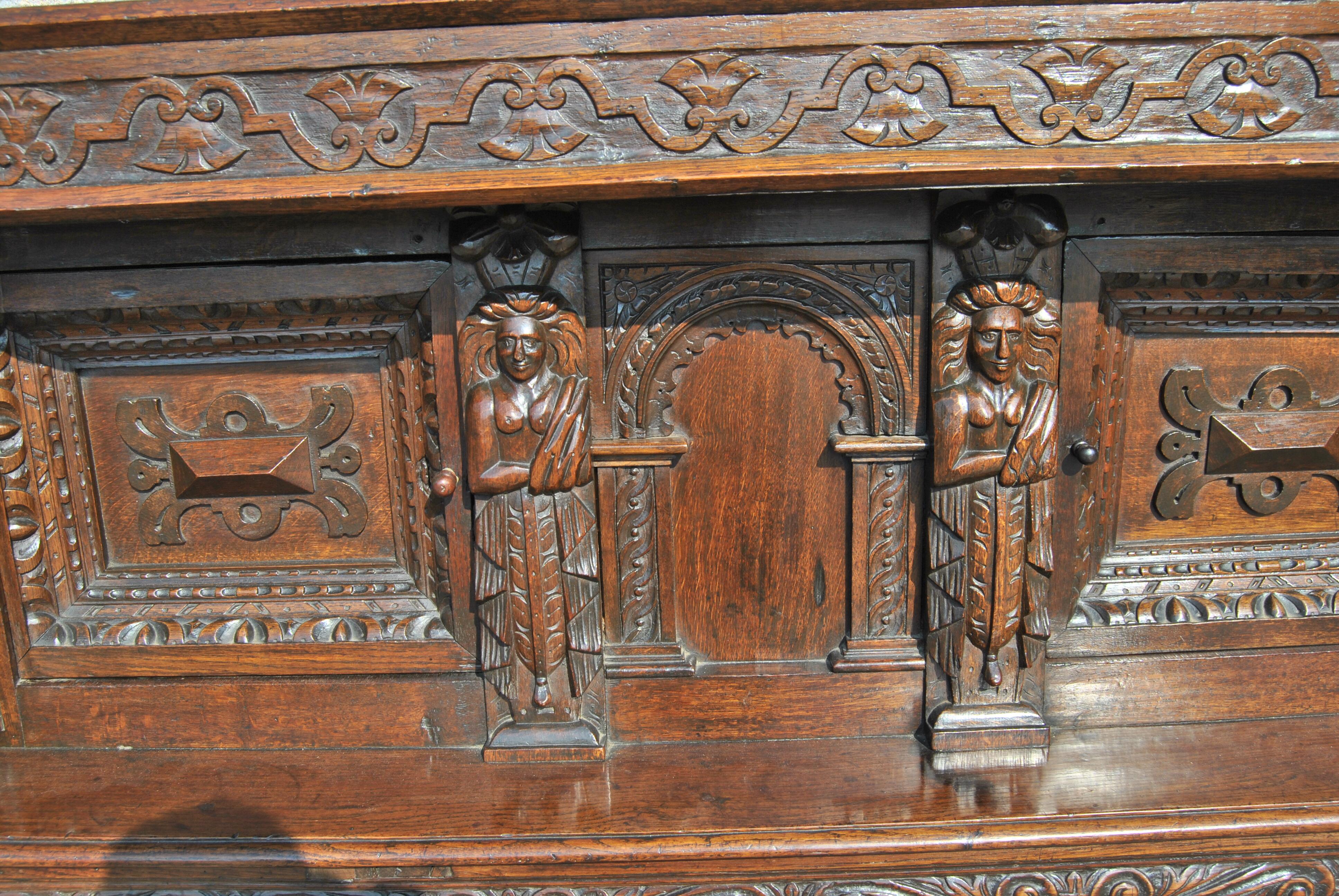 This is a solid oak court cupboard made in England, circa 1725. The top of the piece has a chamfered edge. There is a carved Corinthian topped column on the right and left front. There are 2 doors with deeply recessed hand carving. There is an Atlas