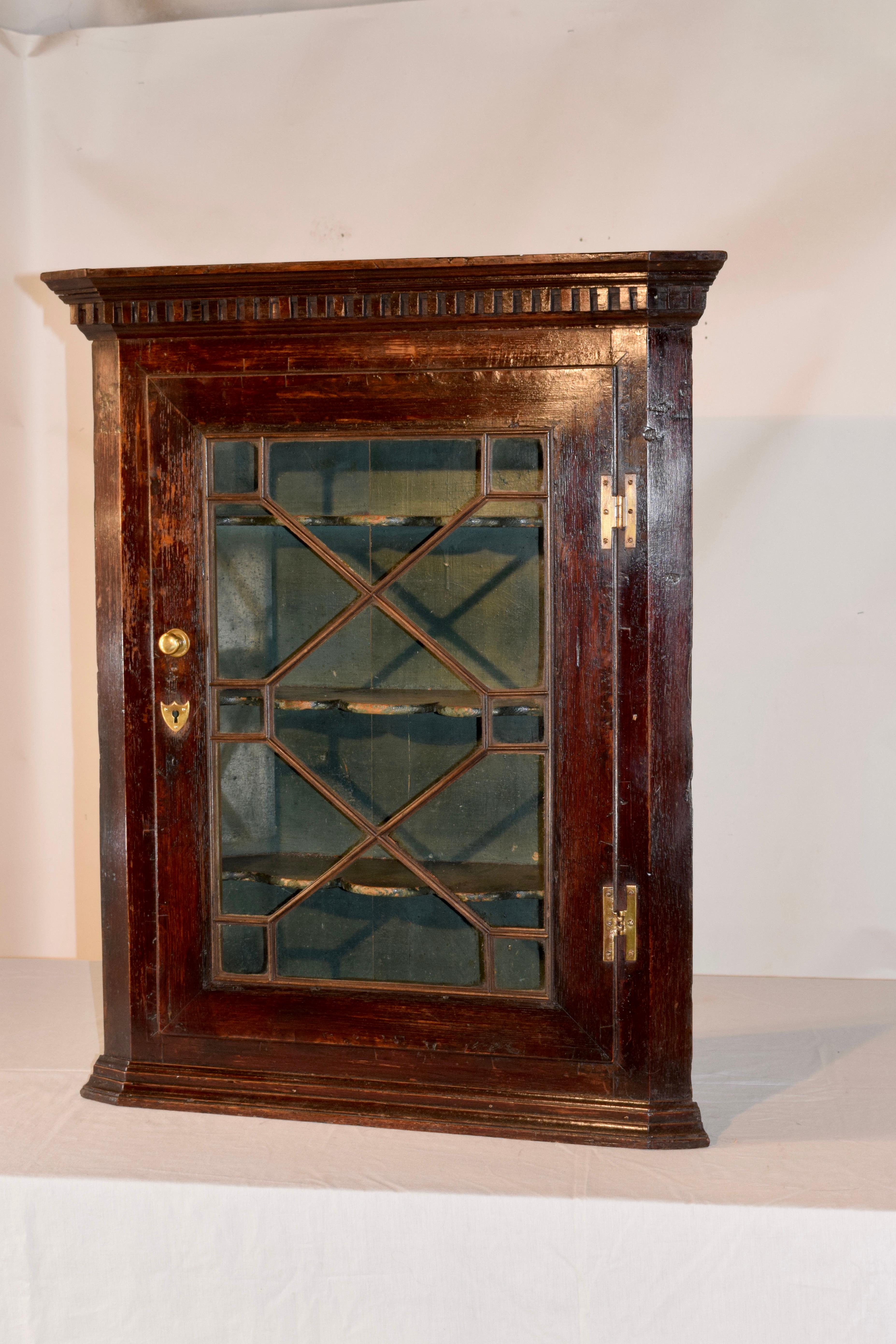 18th century wall cupboard from England made from oak. The piece has crown molding over dentil molding at the top, following down to a single door, which is glazed and opens to reveal three scalloped shelves. The interior is in what appears to be